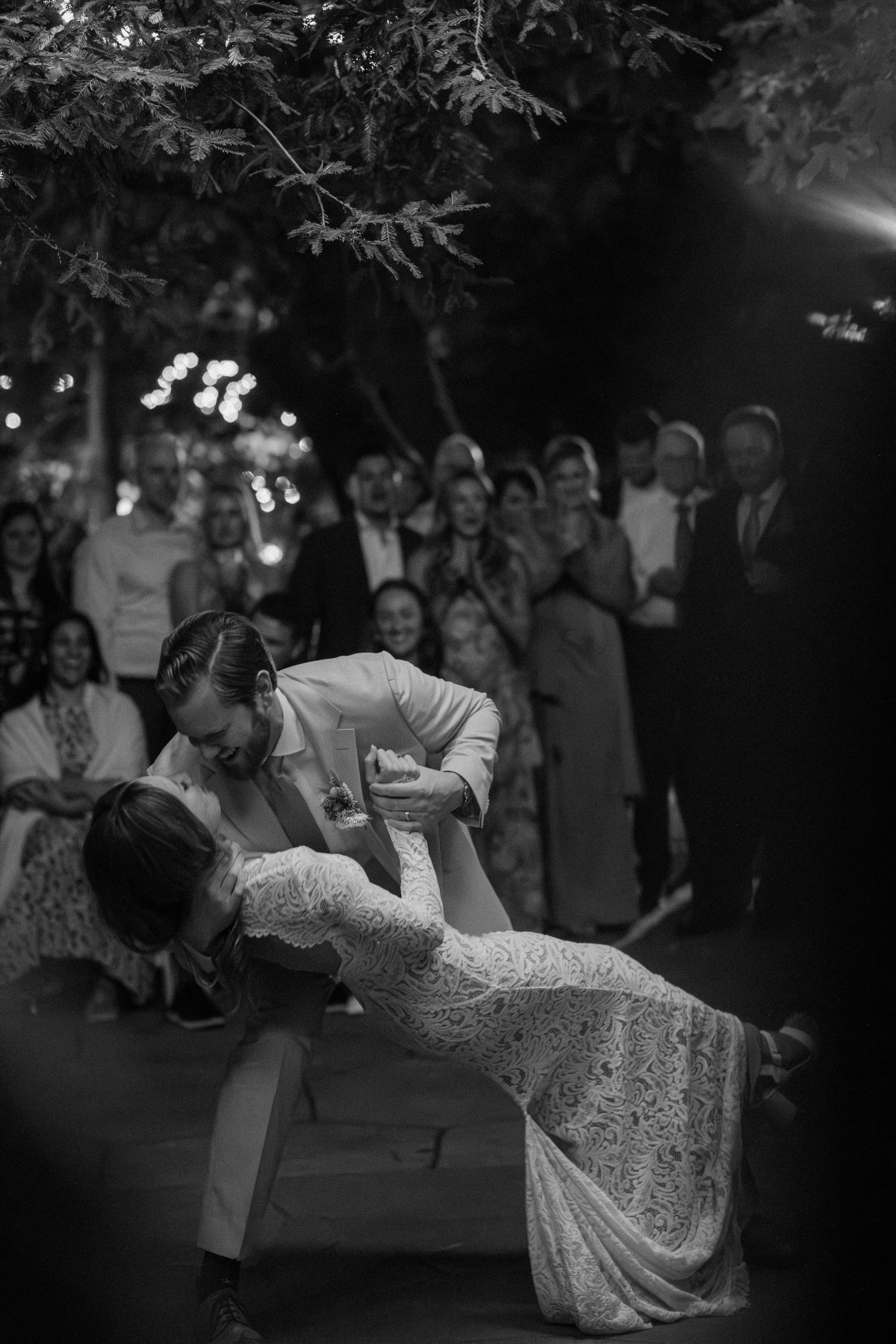 Black and white photo of groom dipping bride during first dance surrounded by wedding guests outdoors.