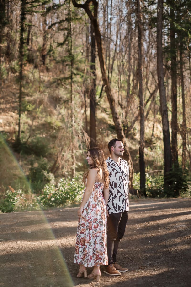 Couple standing together on a sunlit forest path, smiling and enjoying a romantic outdoor moment.