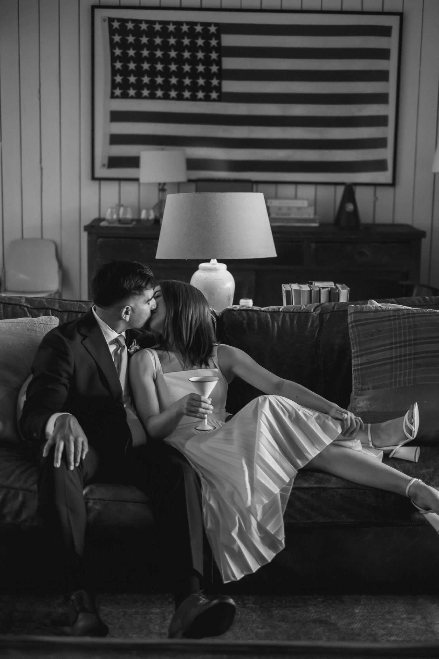 Black and white photo of couple kissing on couch with American flag on wall behind them.