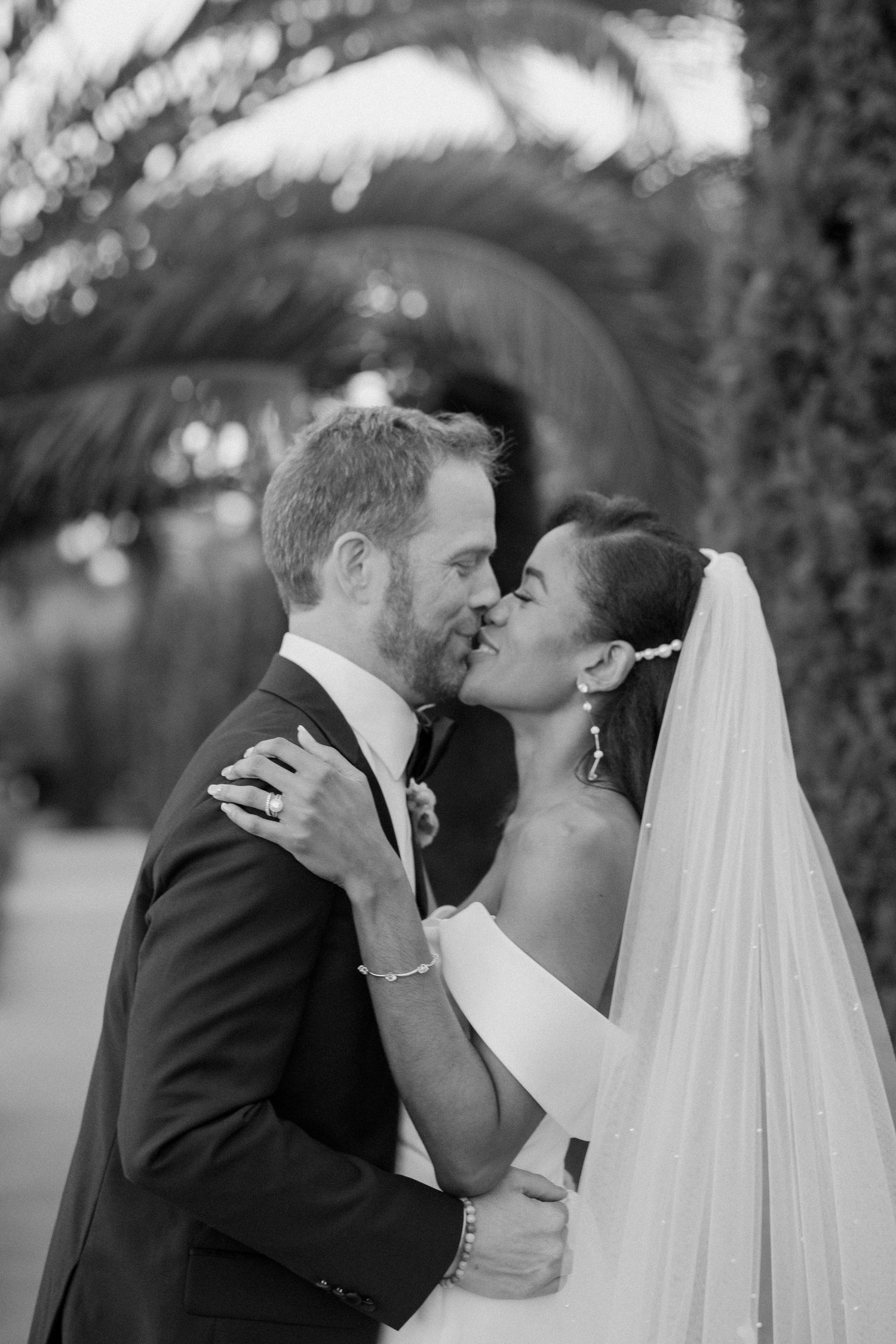 Black and white portrait of bride and groom sharing a kiss under lush palm trees.