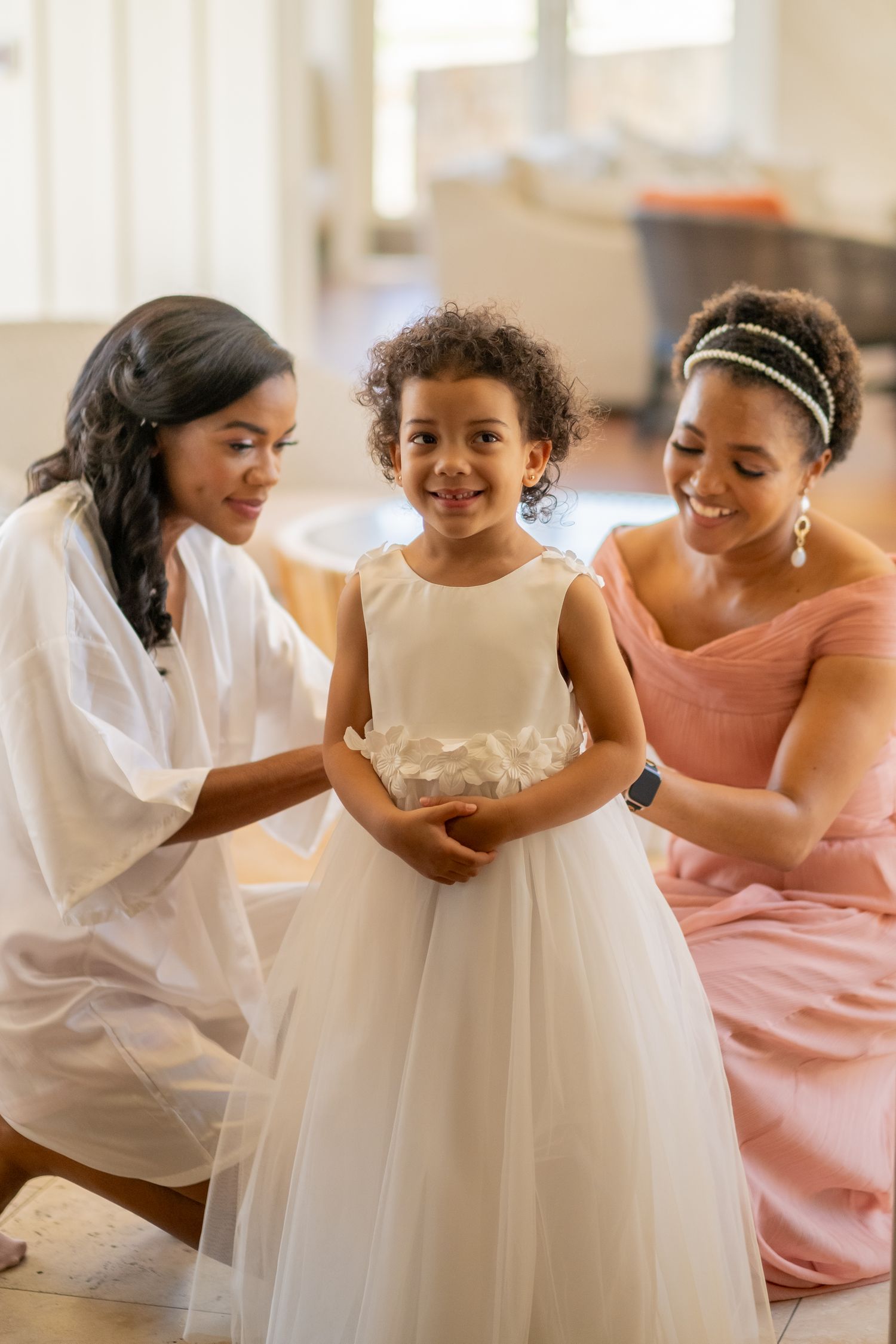 Bride in white robe and bridesmaid in pink dress help a young flower girl in a white tulle gown get ready.