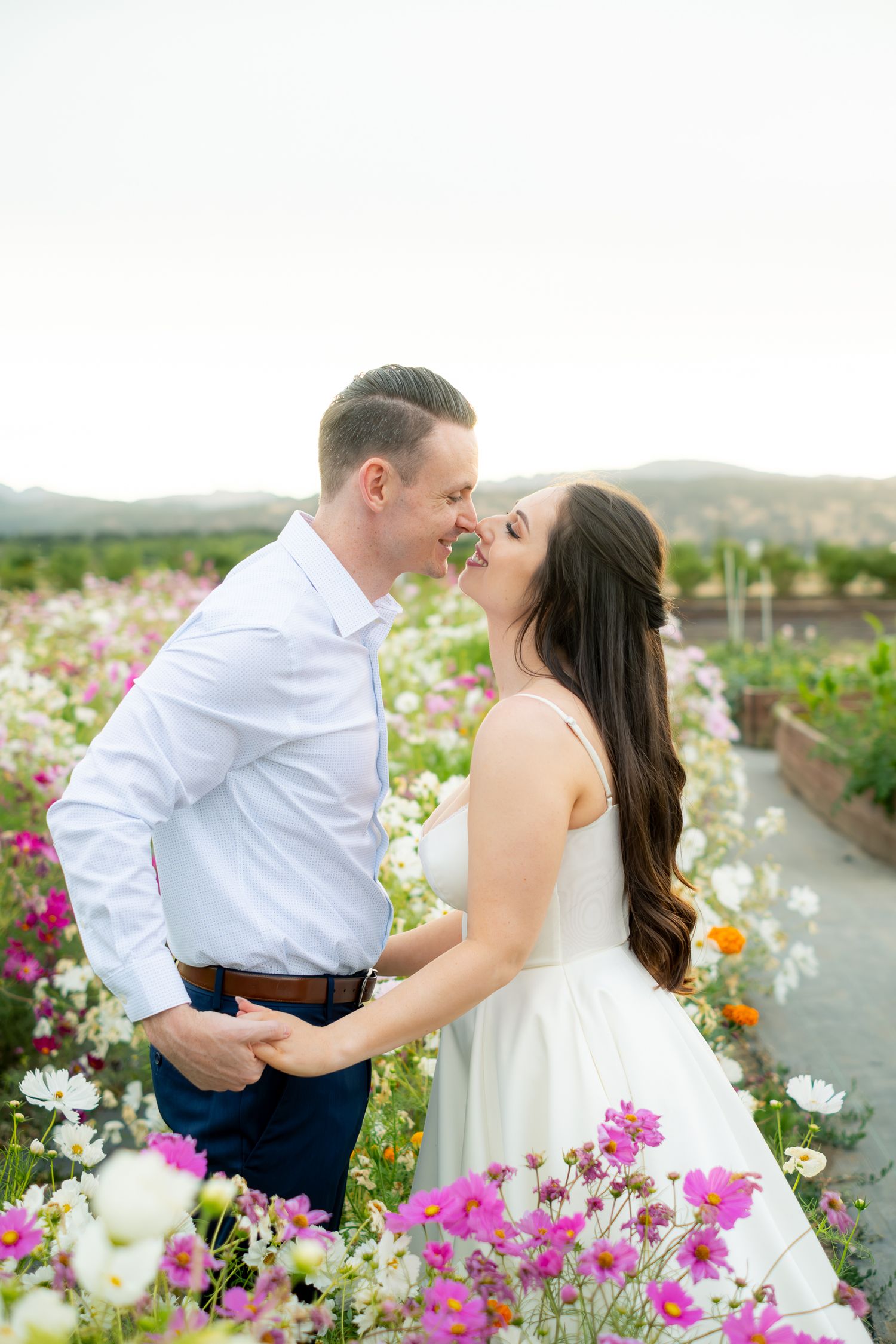 Romantic couple about to kiss surrounded by pink and white wildflowers with mountains in the background.