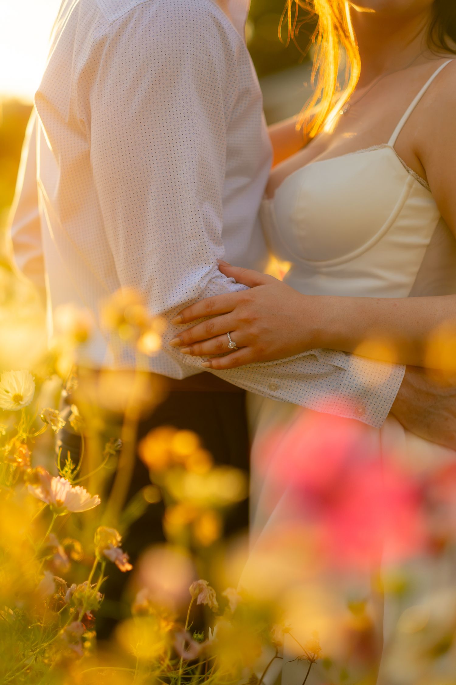 Couple embracing in a sunlit flower field, woman wearing engagement ring, warm golden light glowing around them.