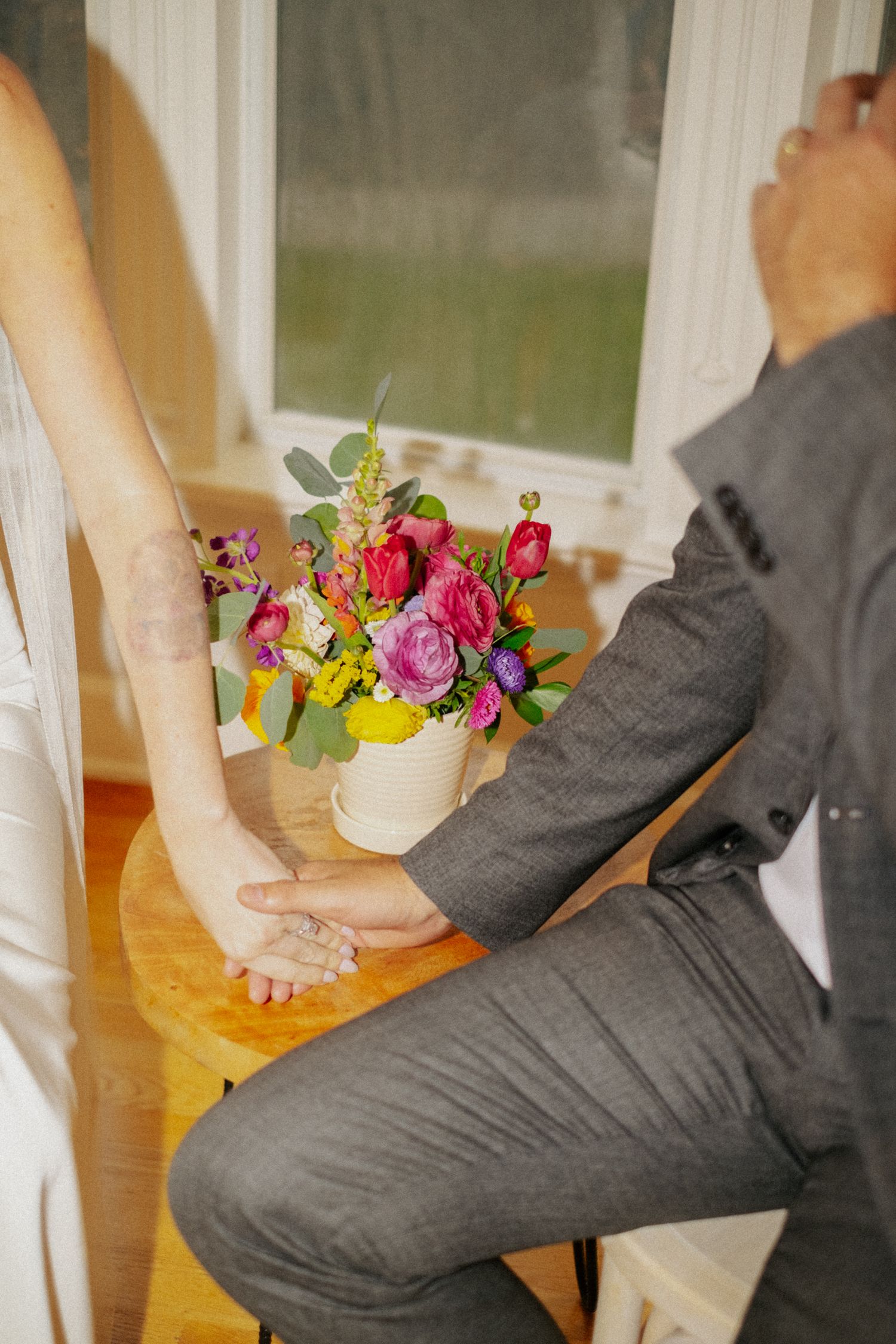 Bride and groom hold hands beside a vibrant colorful floral bouquet with pink, purple, and yellow flowers during wedding.