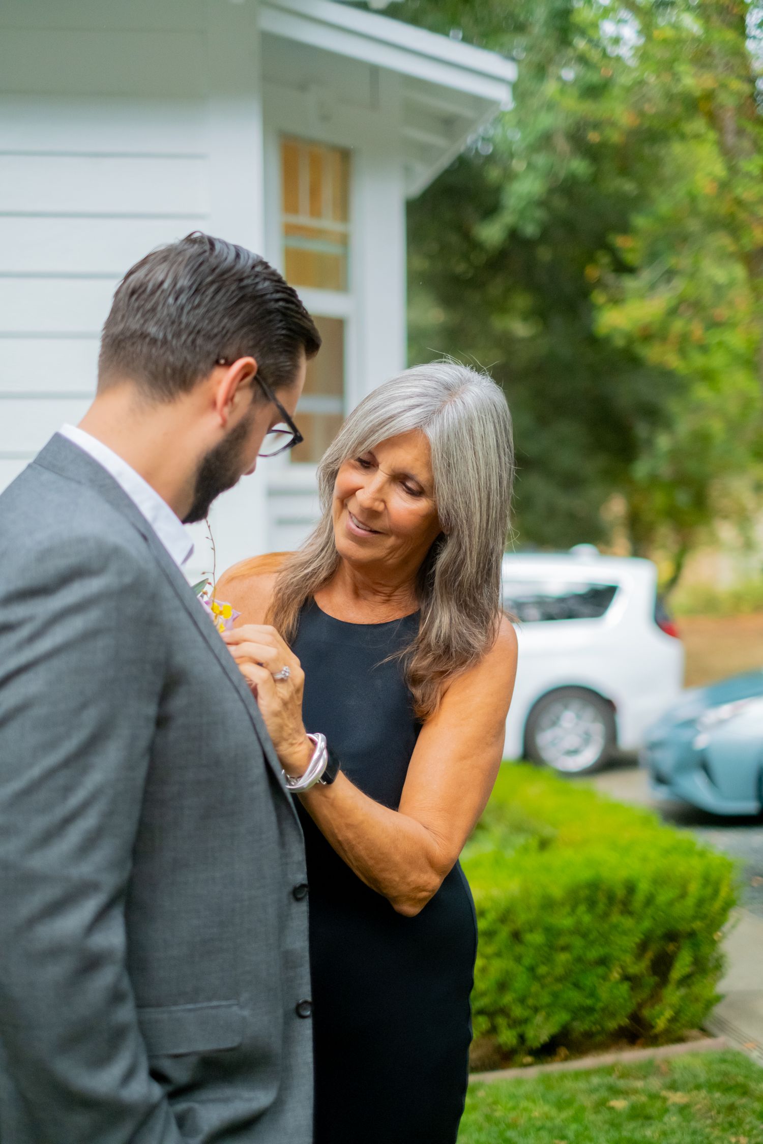 Older woman in black dress pinning boutonniere on man in gray suit outdoors near a white house.