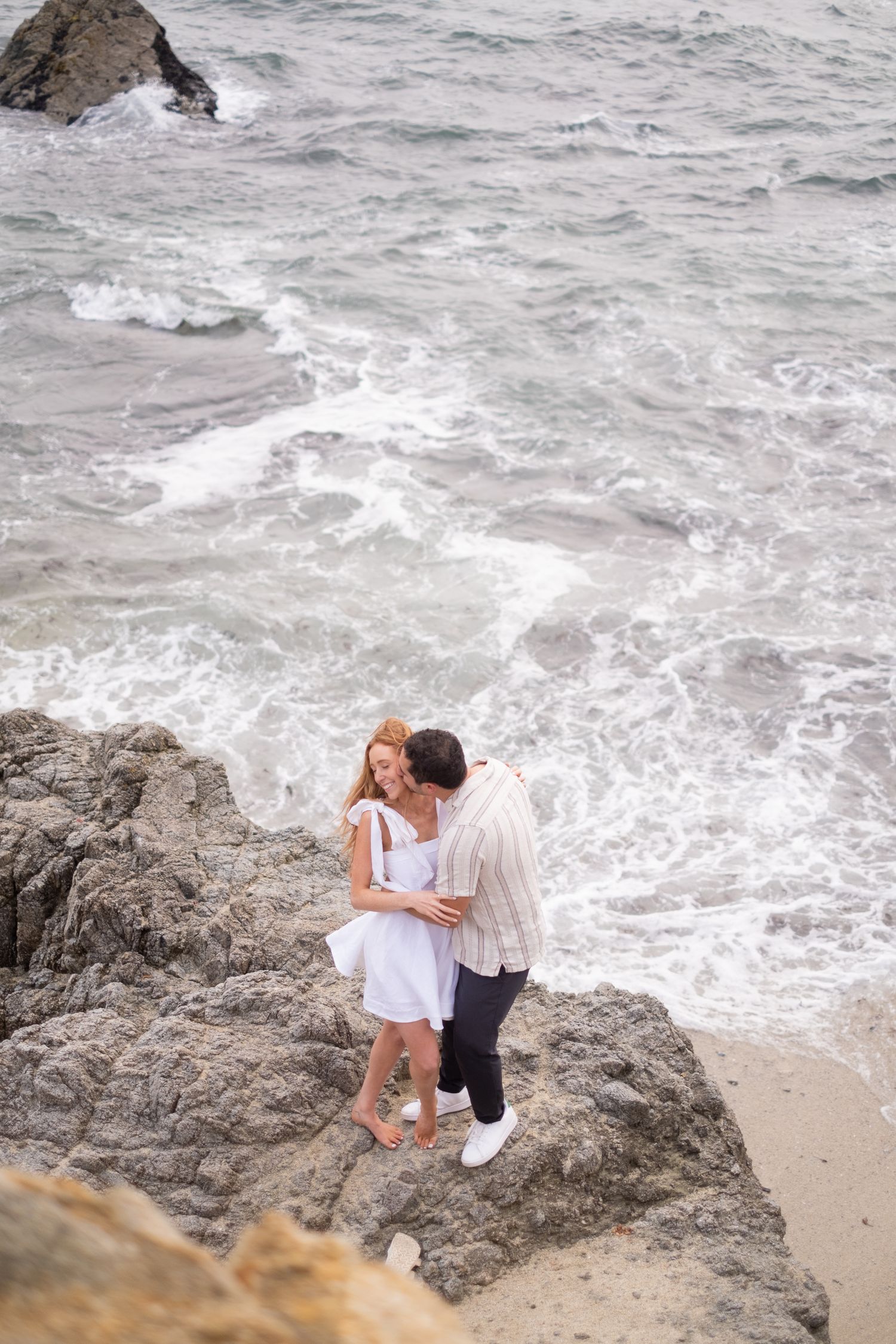Couple embracing on coastal rocks with crashing ocean waves visible below them.