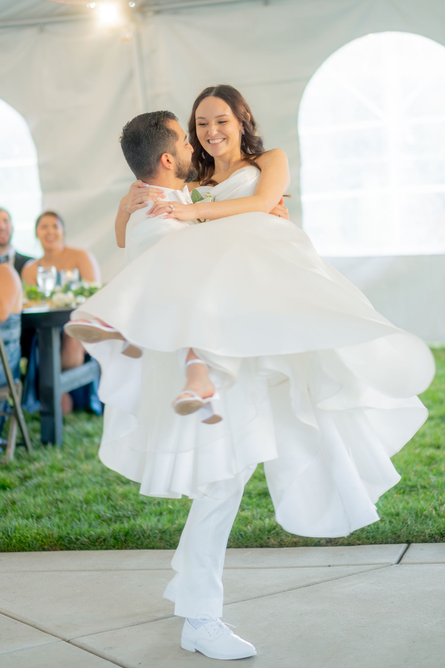 Groom in white suit lifts and spins smiling bride in layered white gown on dance floor.