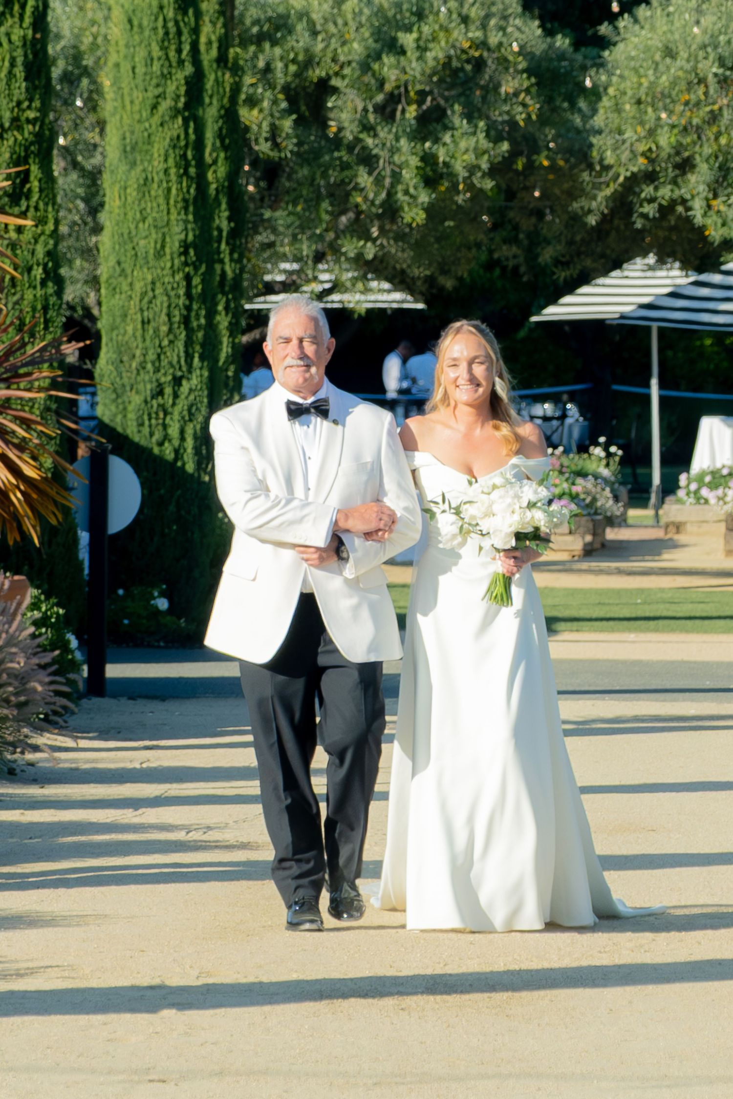 Father escorts smiling bride in white gown down outdoor wedding aisle lined with cypress trees.