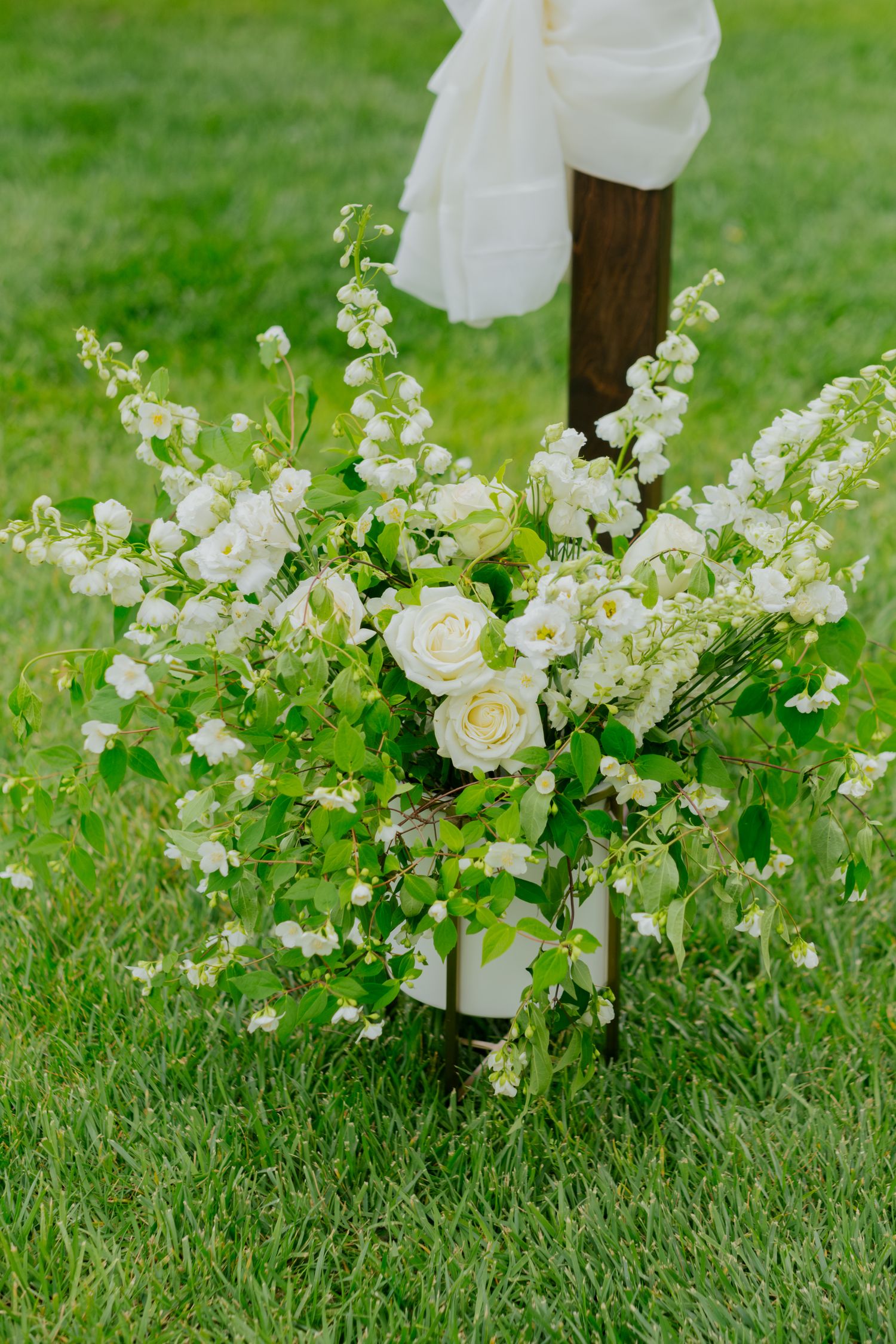 White roses and delicate wildflowers arranged at the base of a rustic wooden wedding ceremony arch post on green grass.