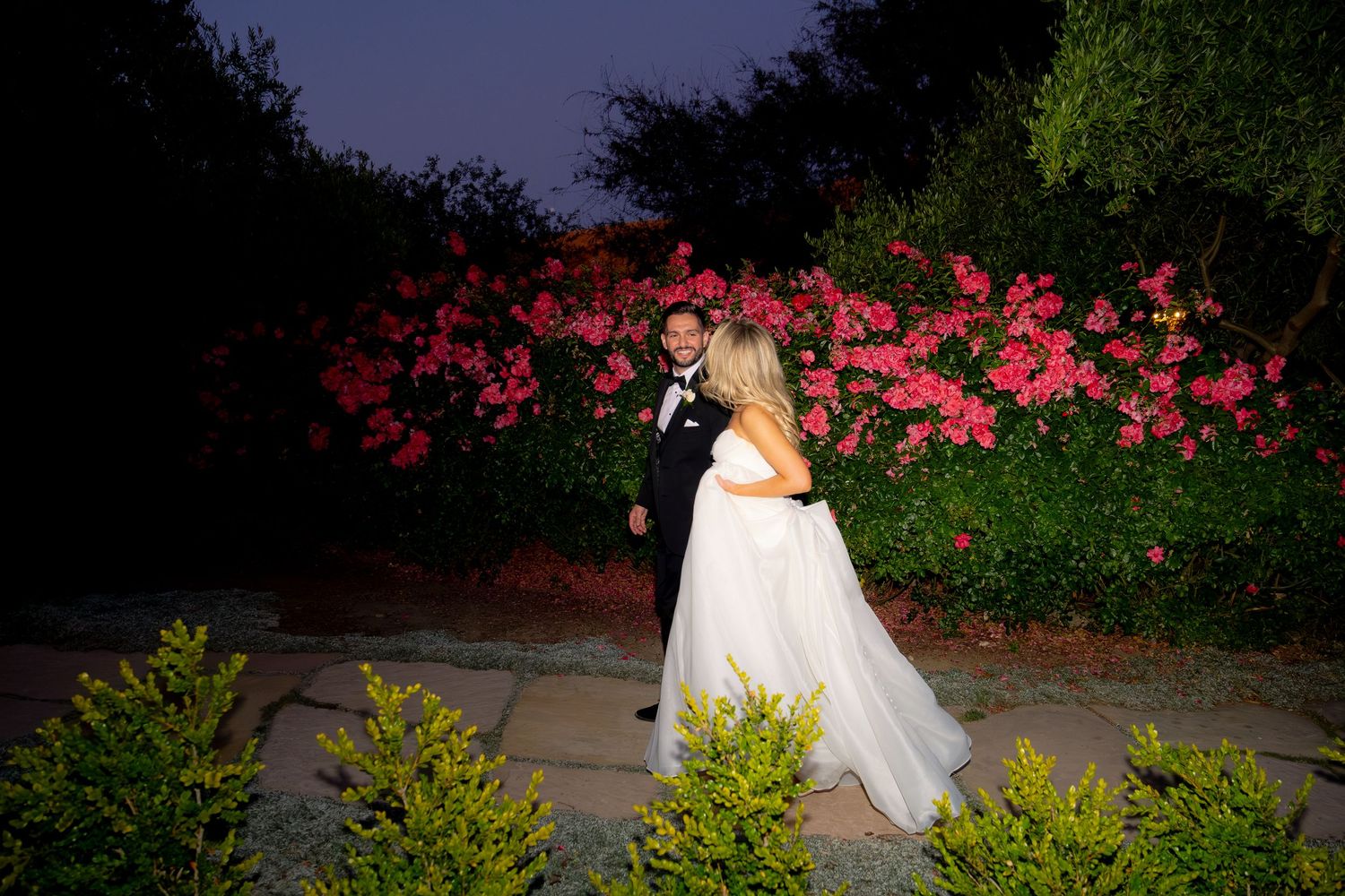 Bride and groom share a romantic moment at night in front of vibrant pink flowering bushes.