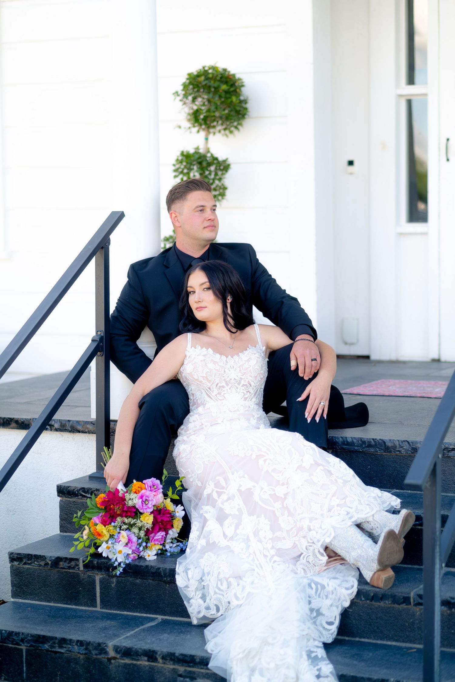 Bride in lace gown and groom in black suit sitting on steps with colorful bouquet at white building wedding portrait.