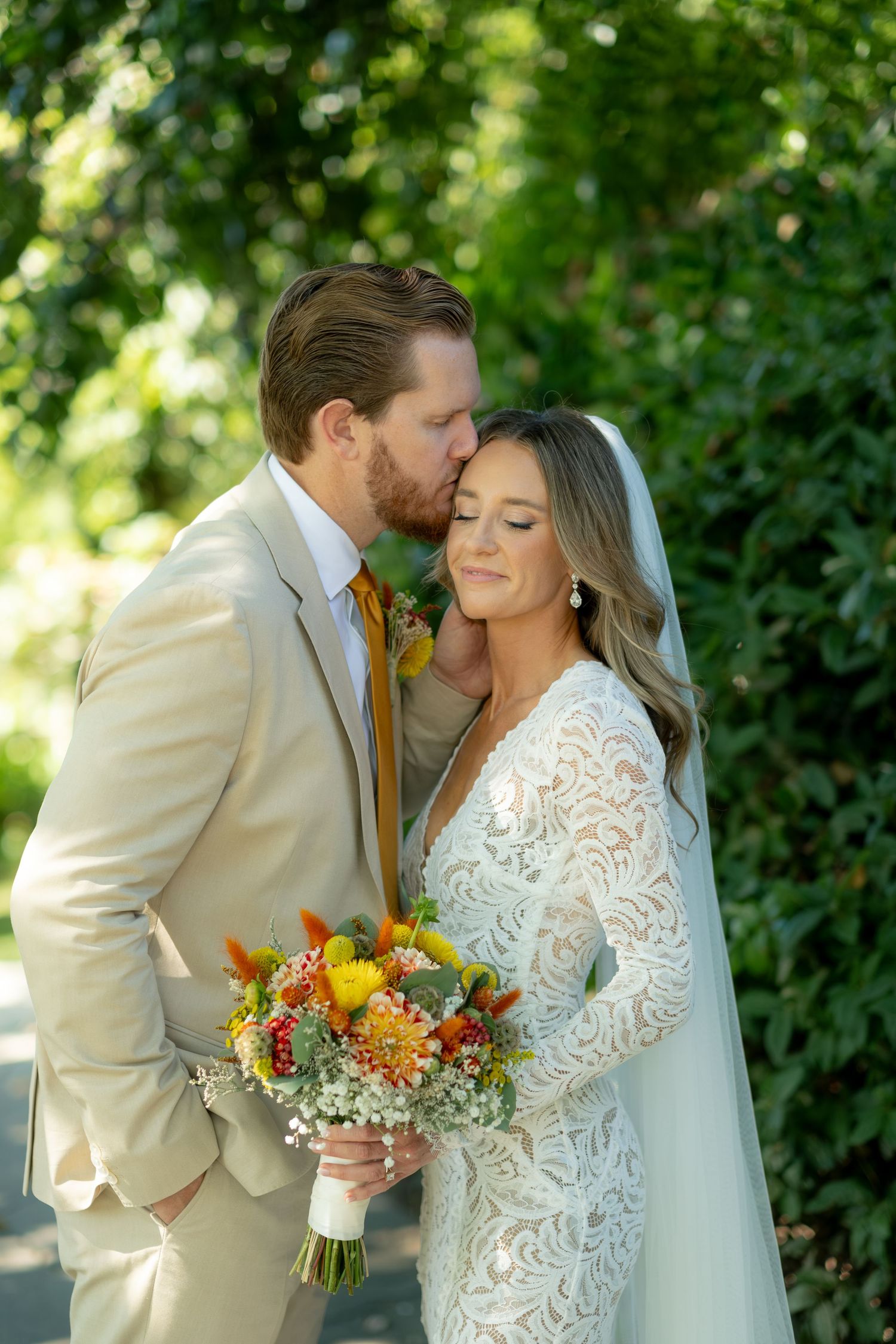Groom in tan suit kissing bride in lace gown holding colorful autumn bouquet outdoors.