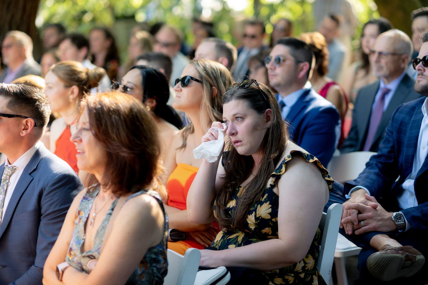 Emotional wedding guests seated outdoors, woman wiping tears during ceremony, crowd in formal attire.
