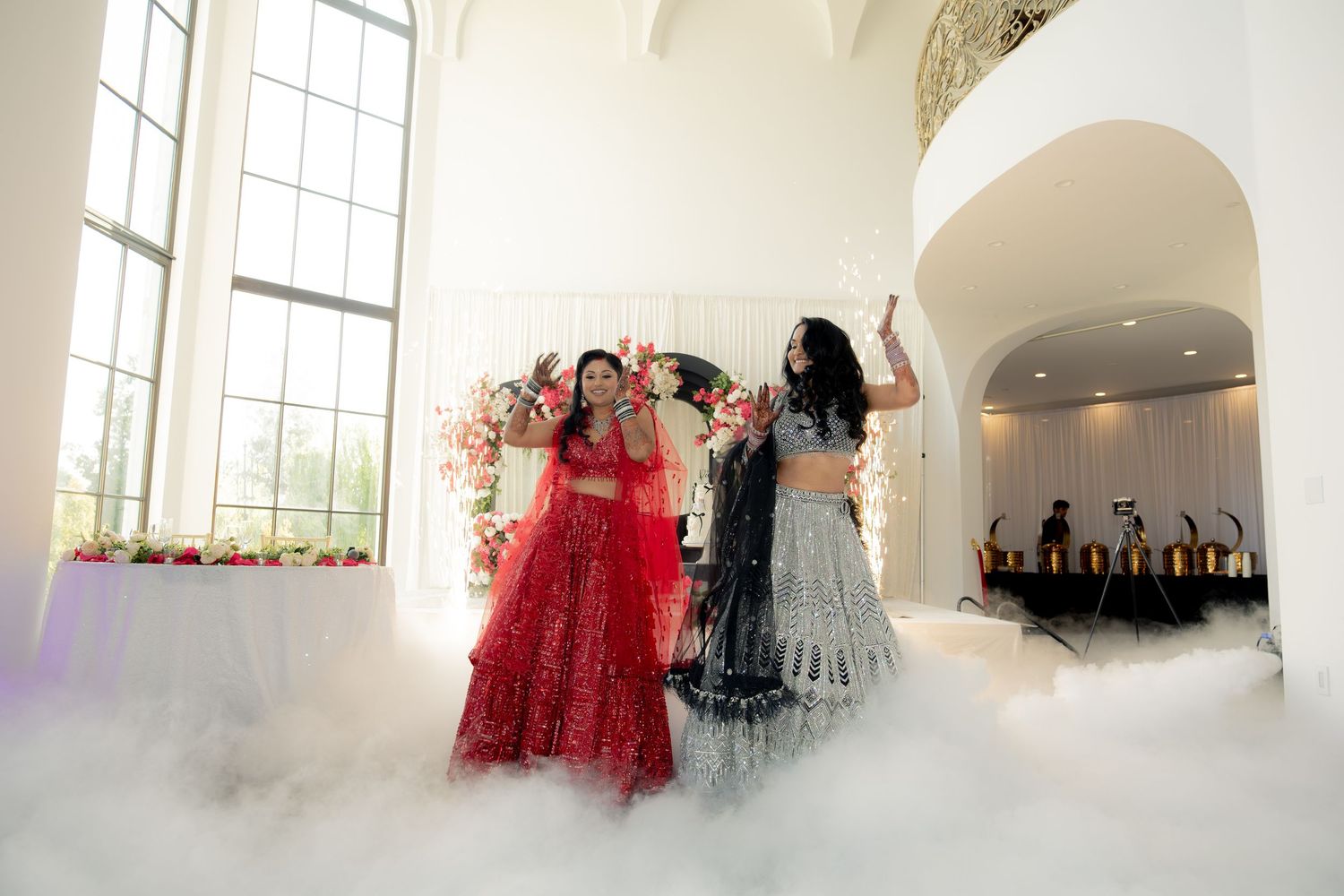 Two women in red and silver lehengas pose dramatically amid dry ice fog at an Indian wedding.