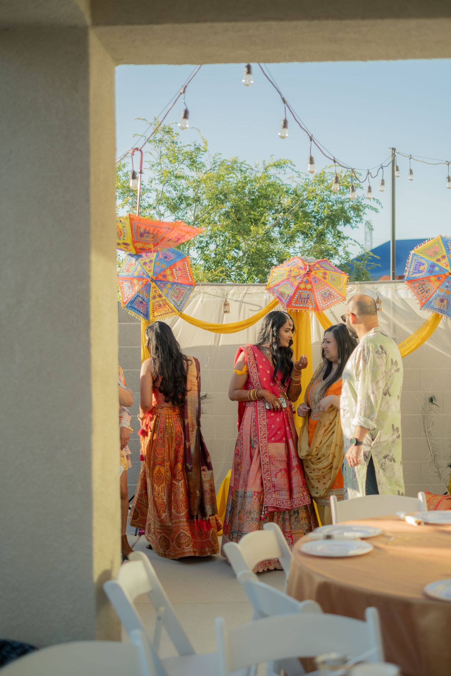 Colorful Indian wedding guests in traditional lehengas gather outdoors under decorative umbrellas and string lights.