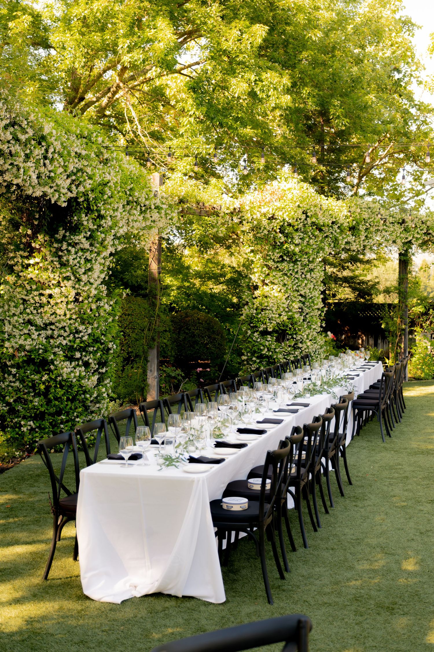 Elegant outdoor wedding reception with a long banquet table set with white linens and black chairs under lush trees.