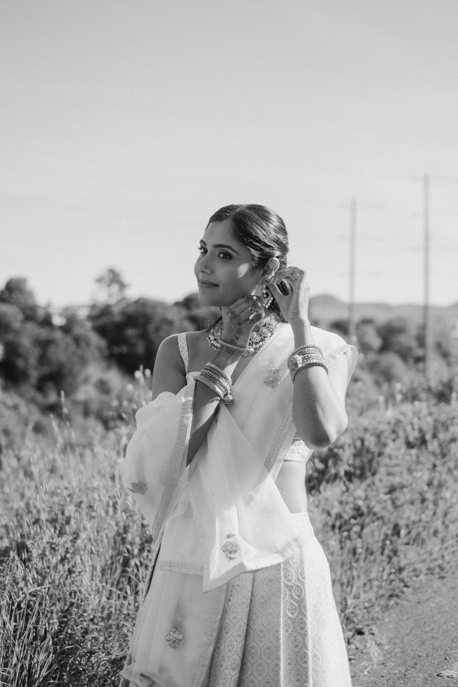 Black and white portrait of a bride in traditional Indian attire adjusting her earring in a field.