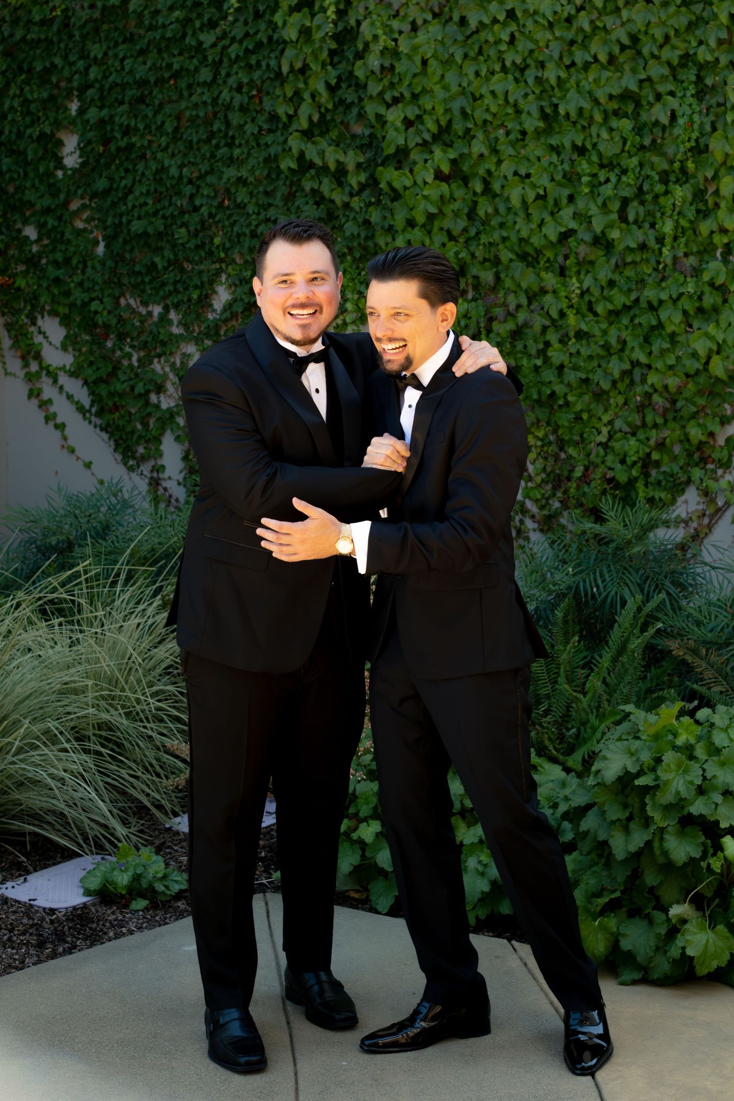 Two grooms in matching black tuxedos embrace and laugh joyfully in front of a green ivy wall.