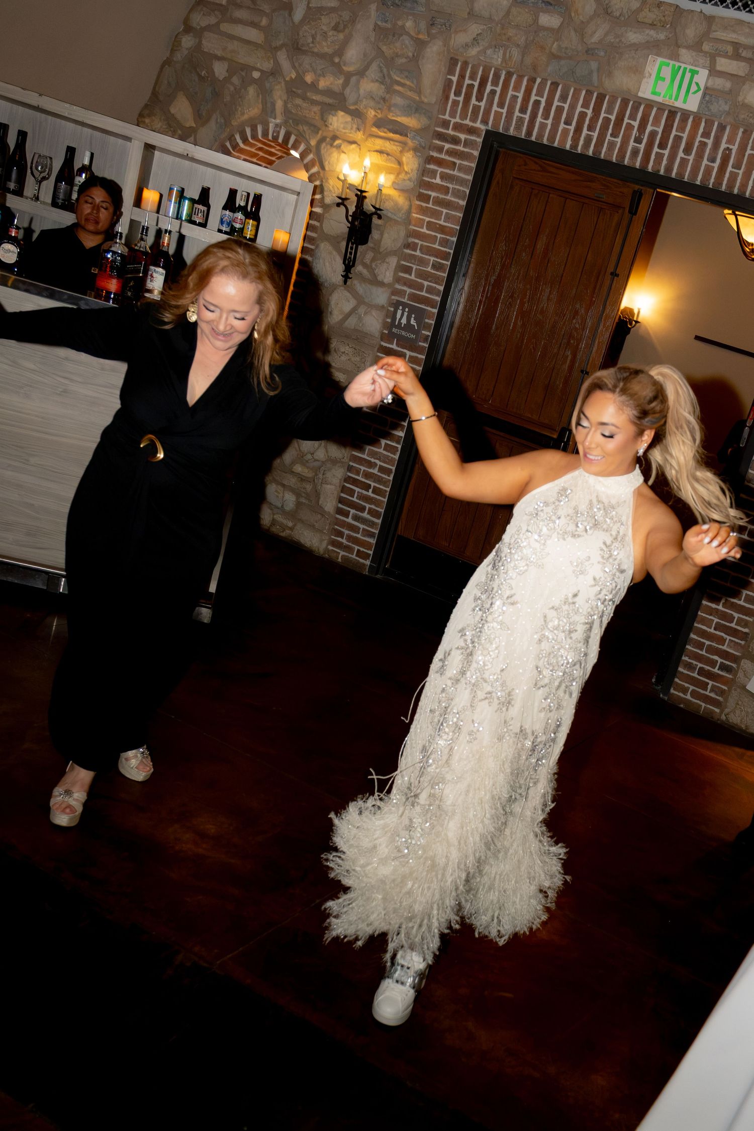 Bride in white feathered gown dancing with guest in black outfit at wedding reception on dance floor.