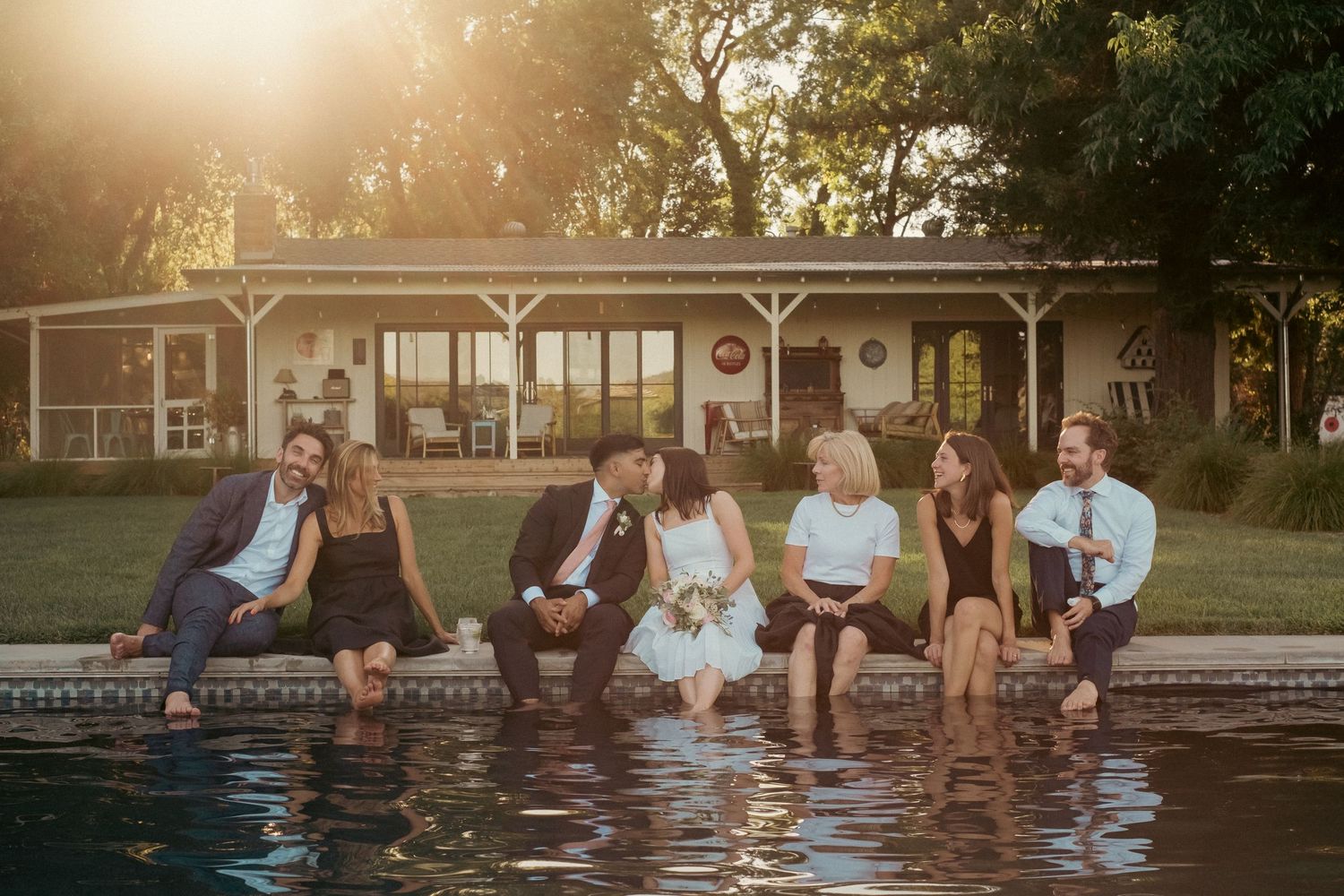 Wedding party sitting poolside with feet in water at sunset during outdoor reception celebration.