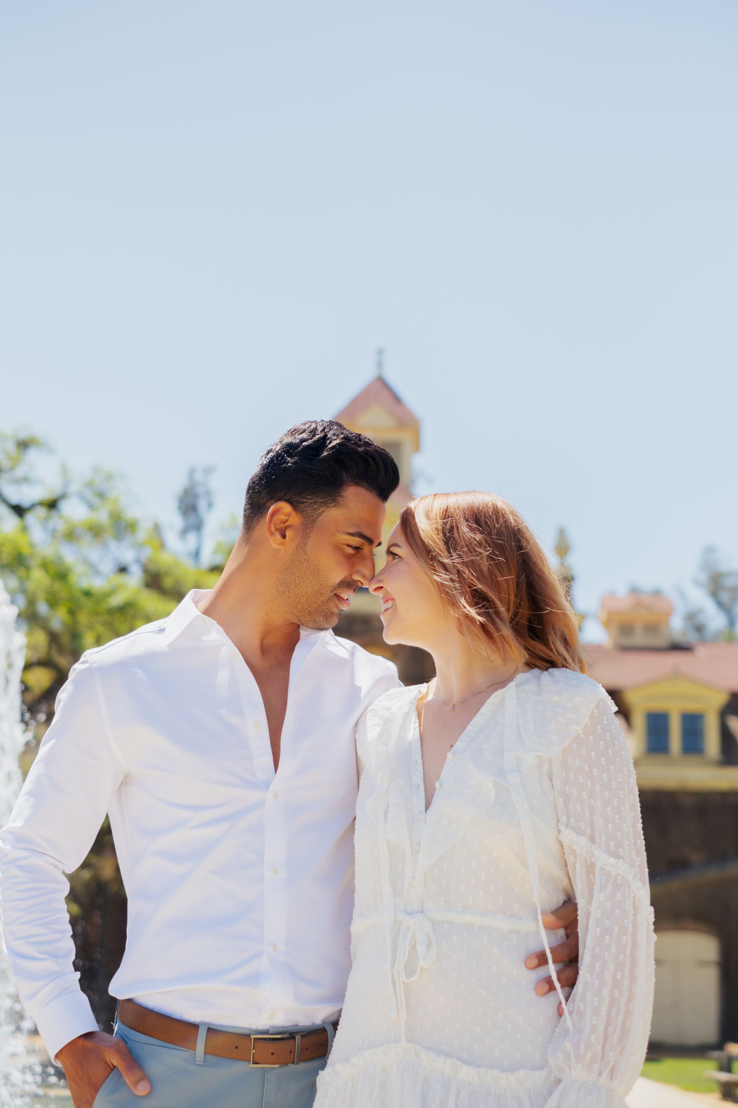Couple in white outfits sharing a tender forehead-to-forehead moment near a fountain on a sunny day.