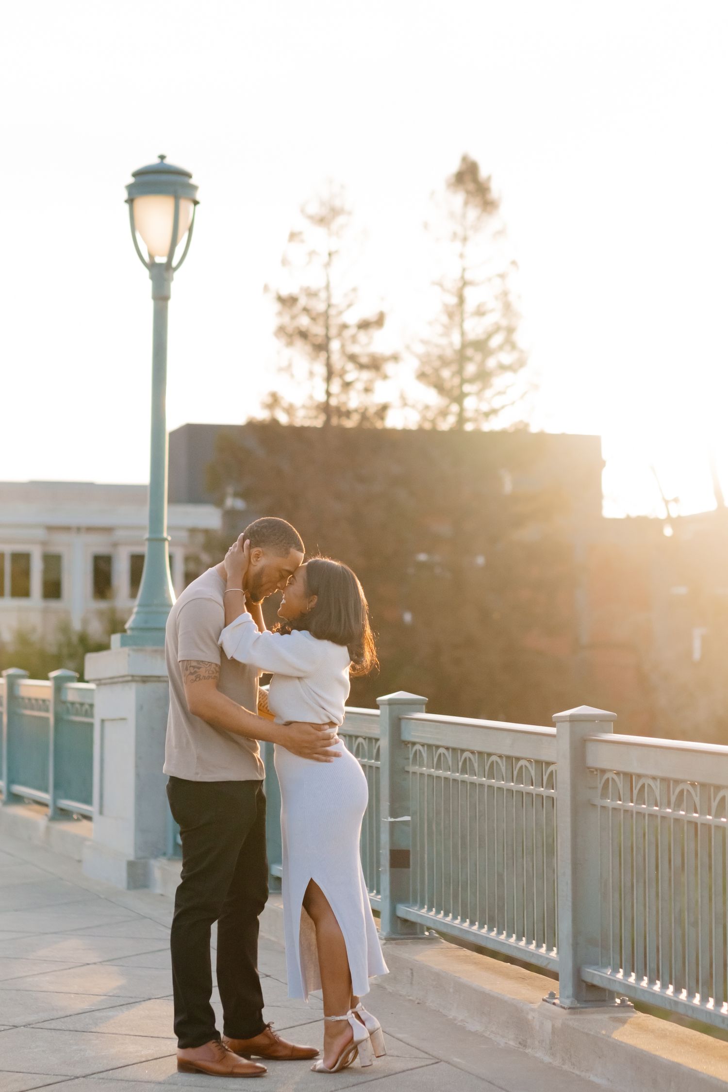 A romantic couple embraces and kisses on a bridge at golden hour with warm sunlight behind them.