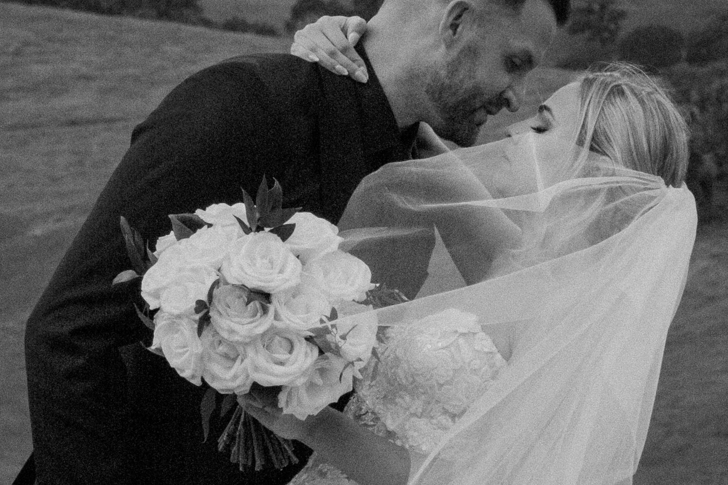 Black and white photo of bride and groom kissing under flowing veil outdoors.