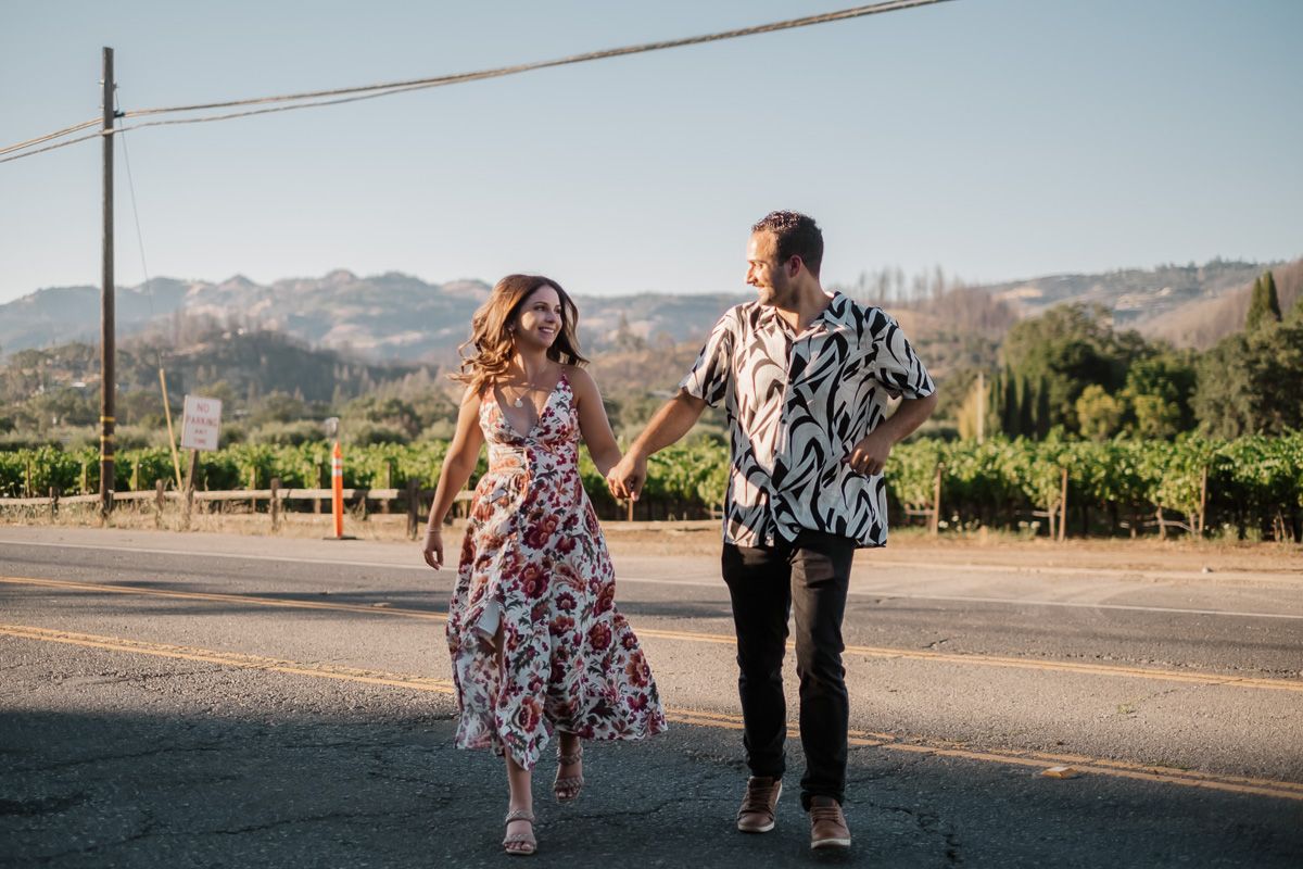 Couple holding hands and walking along a road with lush green vineyard and hills behind them.