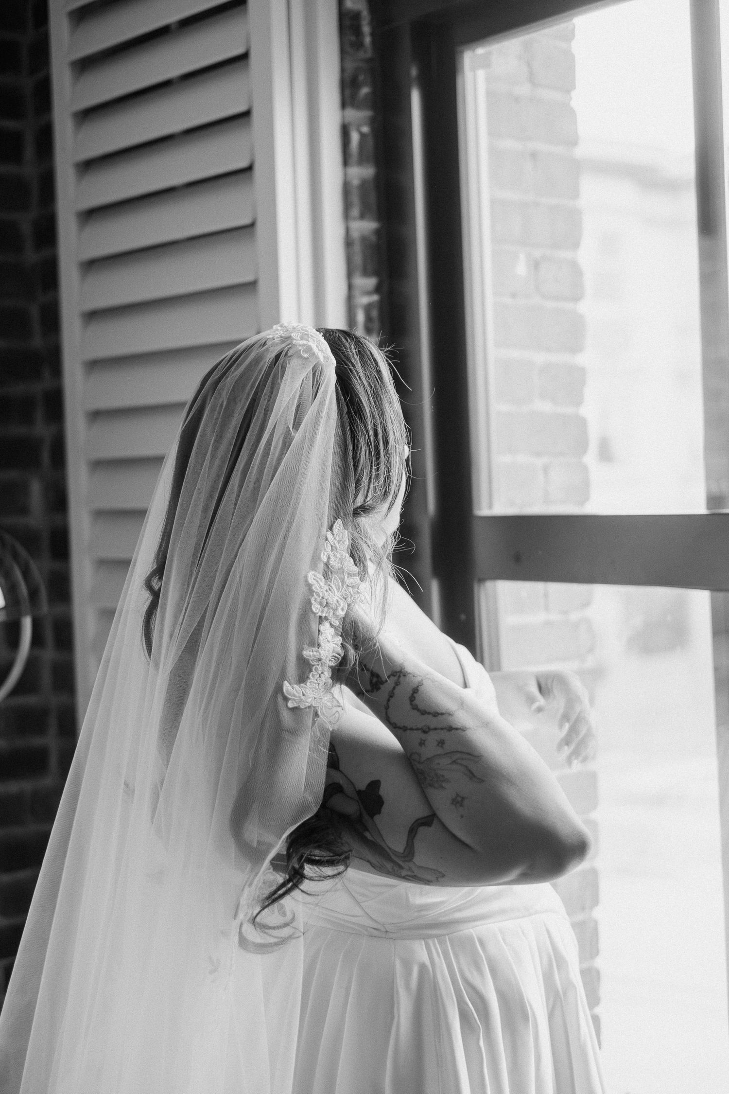 Black and white photo of a tattooed bride in a veil gazing out a window on her wedding day.