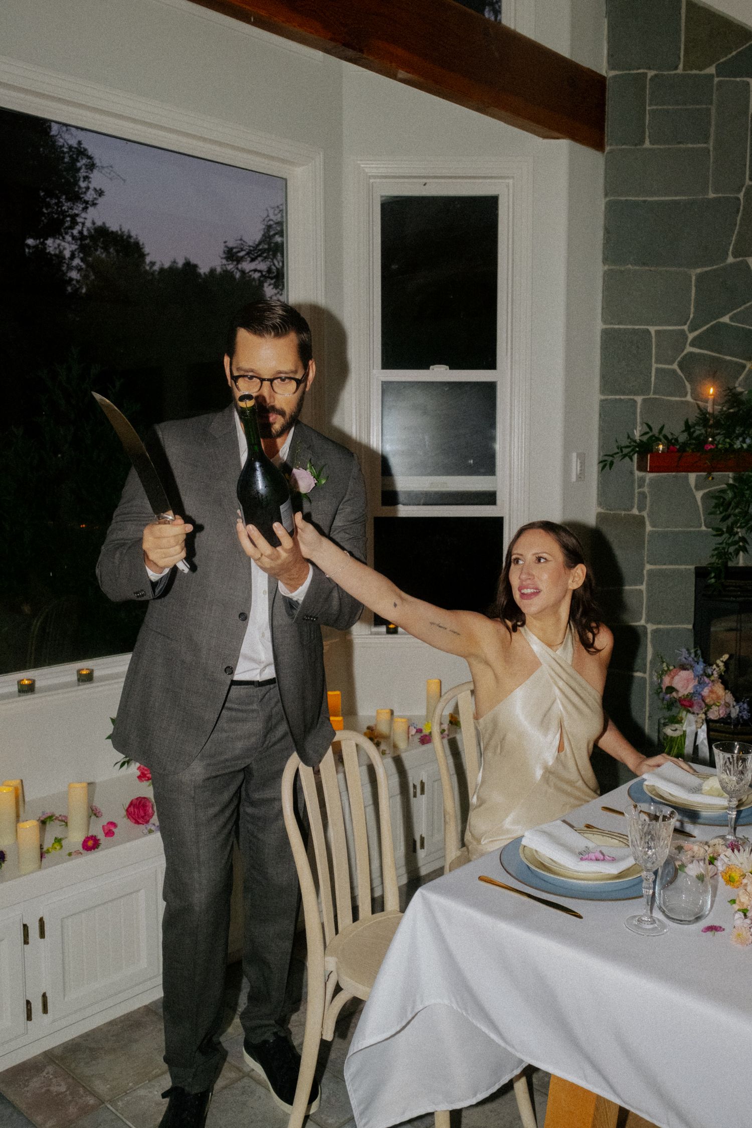Groom in gray suit sabering champagne bottle as bride laughs at elegant indoor wedding reception dinner.
