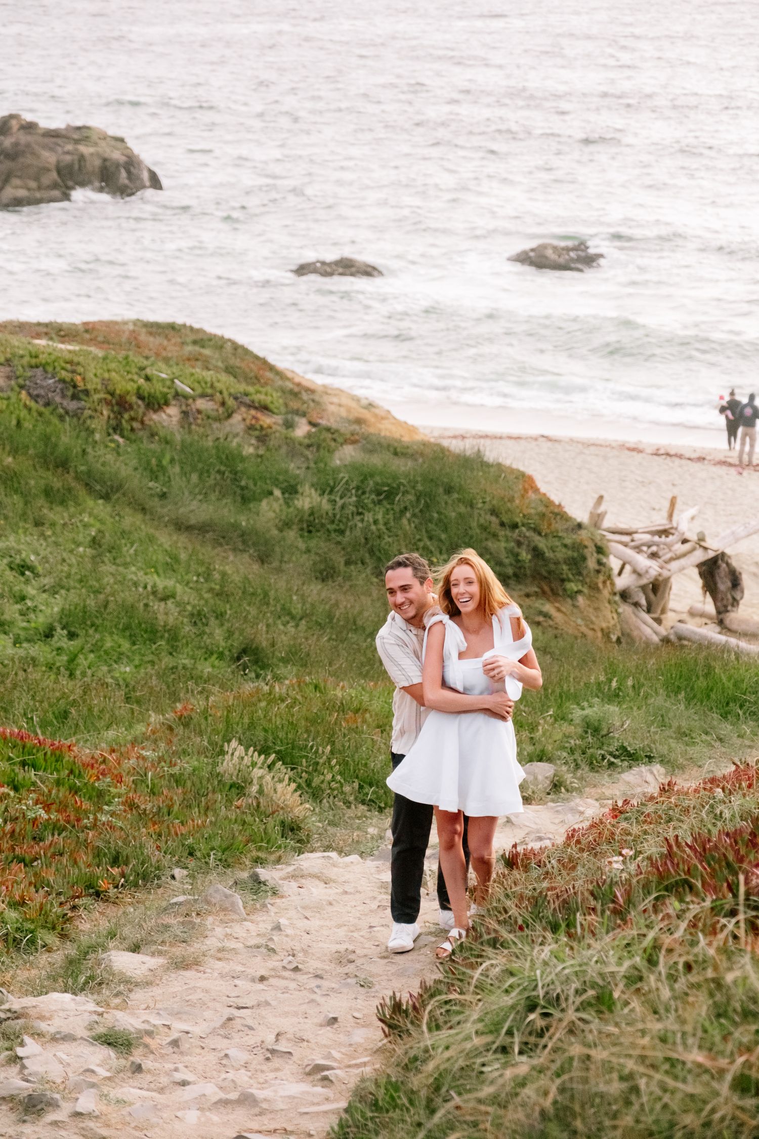 Couple embracing on a coastal cliff path with ocean waves and green hillside in the background.