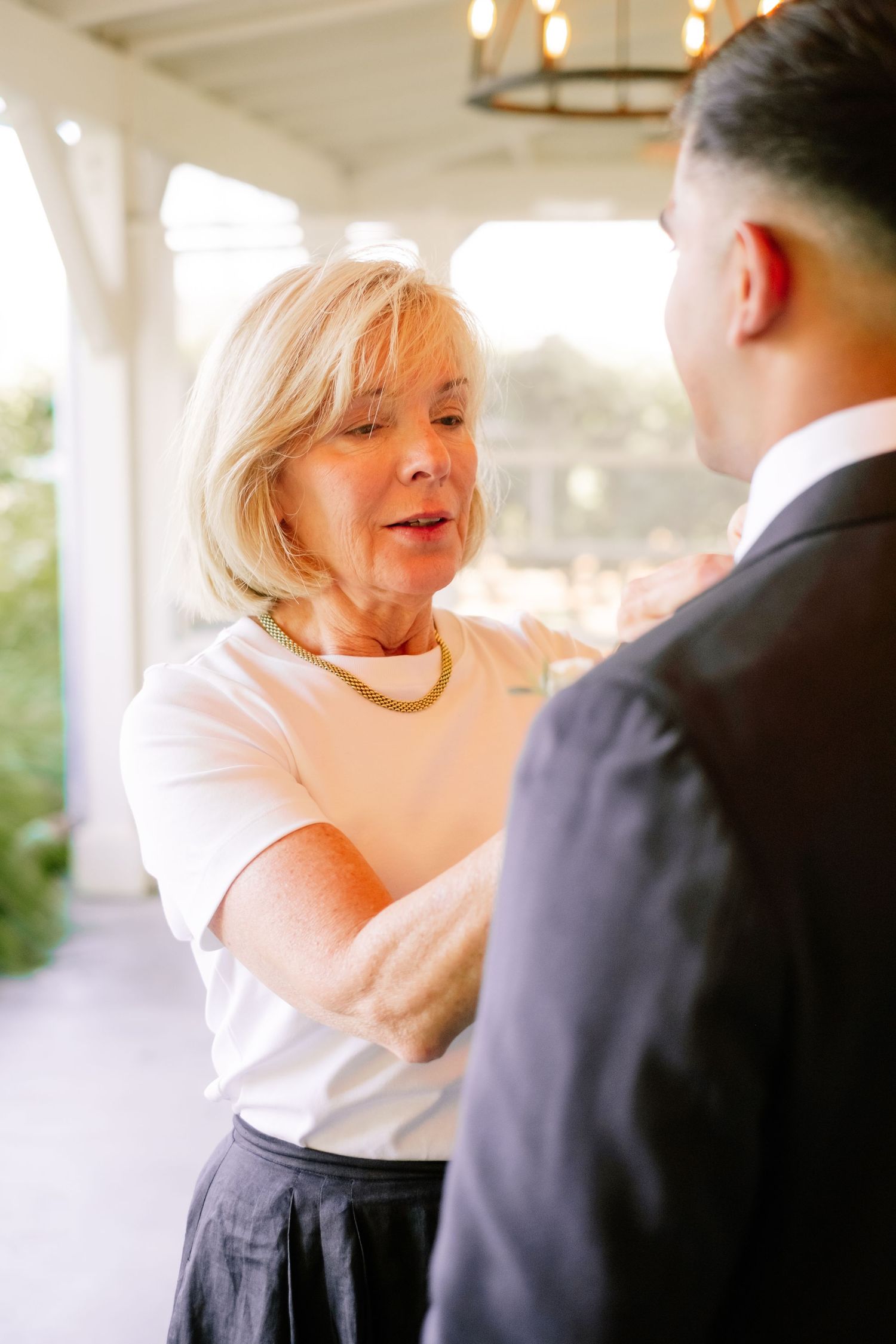 Elegant older woman in white top and gold necklace adjusts man's suit jacket on outdoor porch.