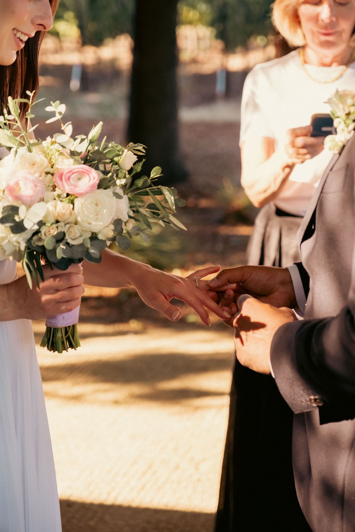 Groom placing wedding ring on bride's finger during outdoor ceremony, bride holding pink and white floral bouquet.