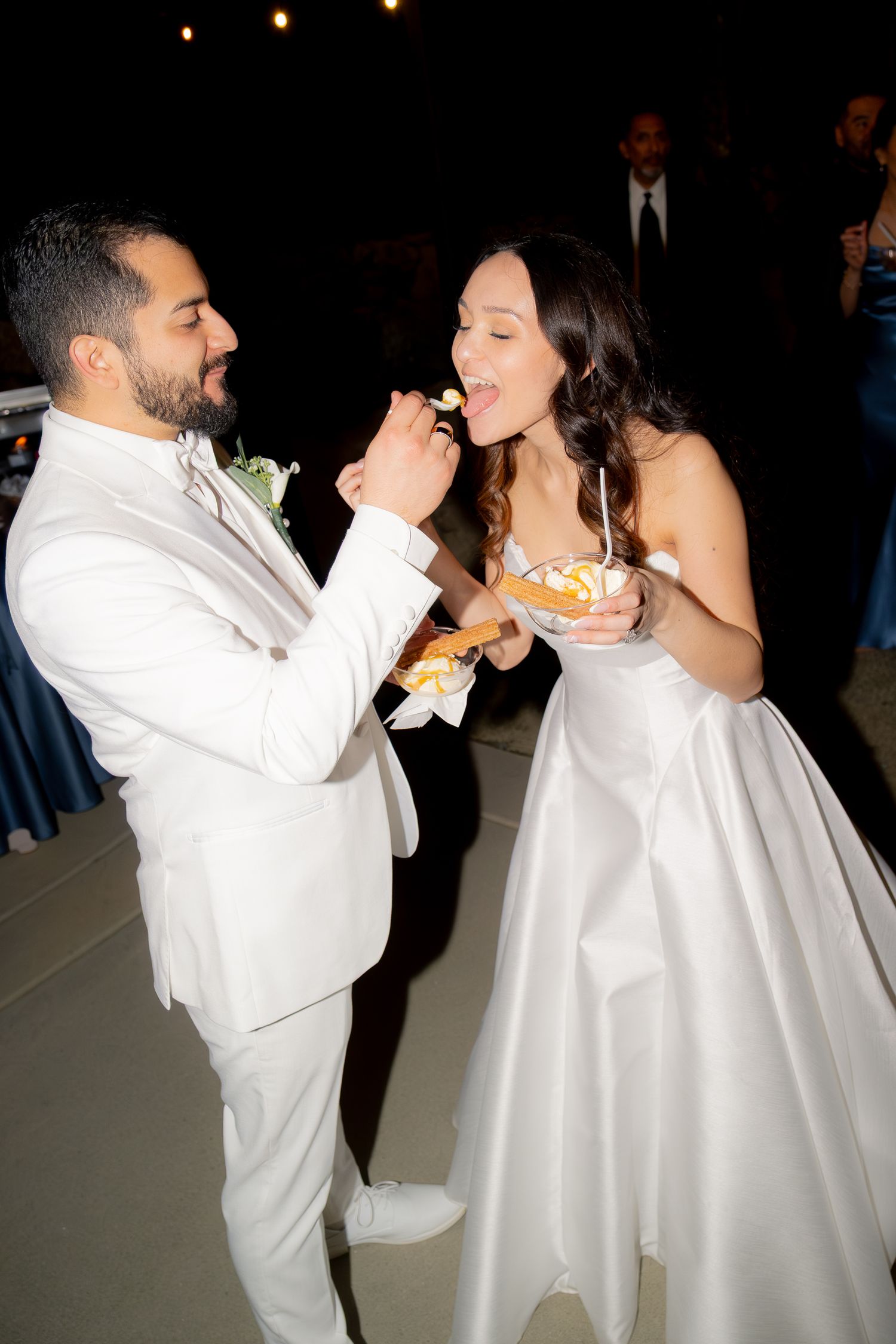 Groom in white suit feeding bride from a dessert bowl at evening wedding reception.