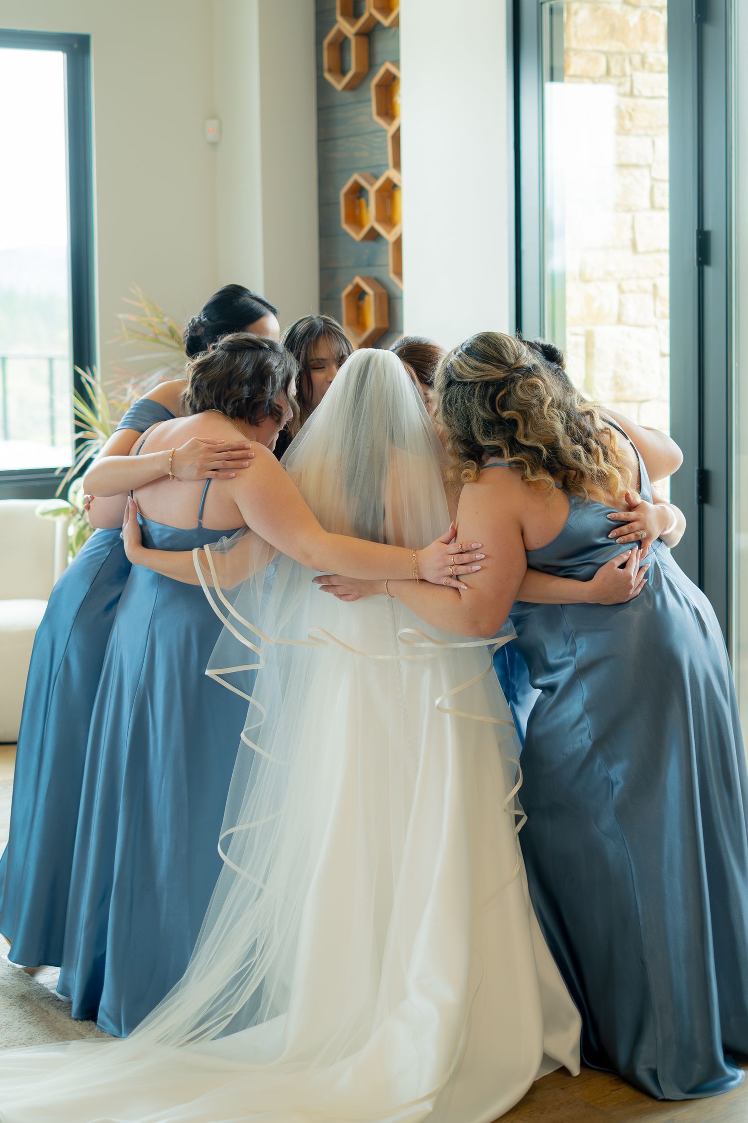 Bride in white gown and veil shares emotional group hug with bridesmaids in blue dresses.