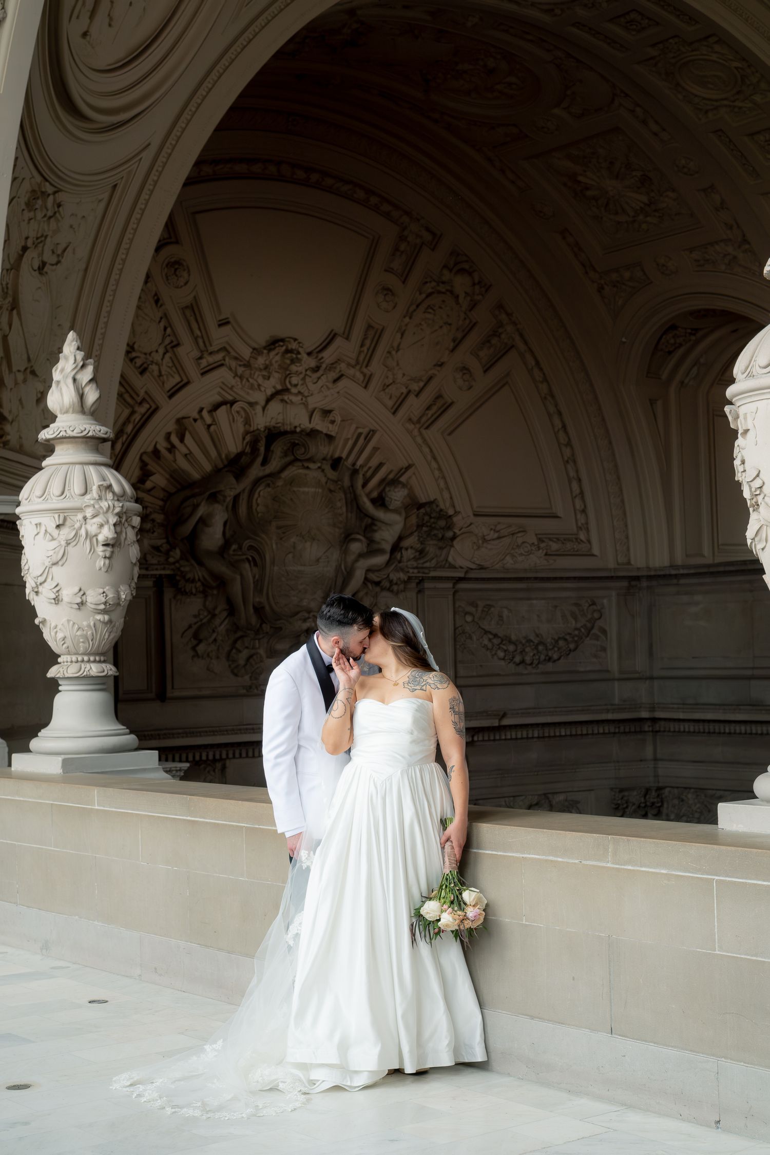 Bride and groom share a kiss under ornate stone arches at a grand wedding venue.
