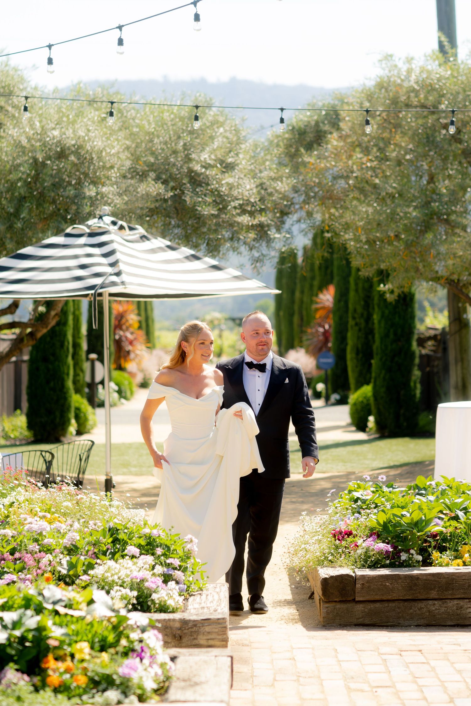 Bride and groom walking through flower-lined garden path with string lights at sunny outdoor wedding.