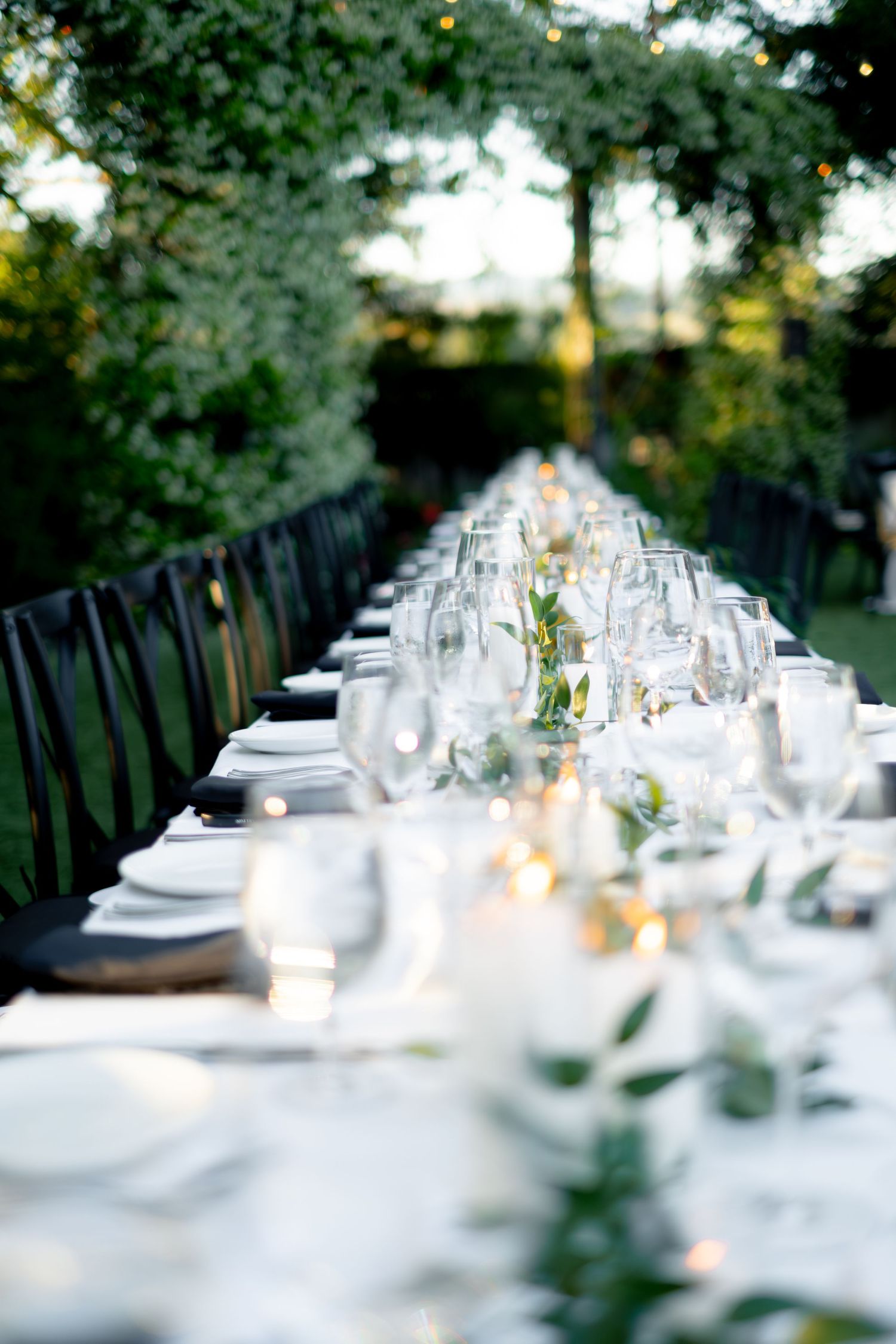 Elegant long outdoor wedding reception table set with white linens, candles, and greenery centerpieces.
