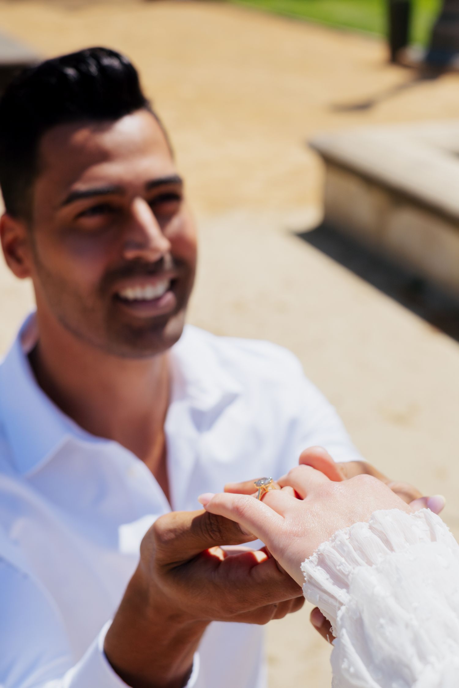 Smiling man in white shirt placing an engagement ring on his partners finger during a proposal.