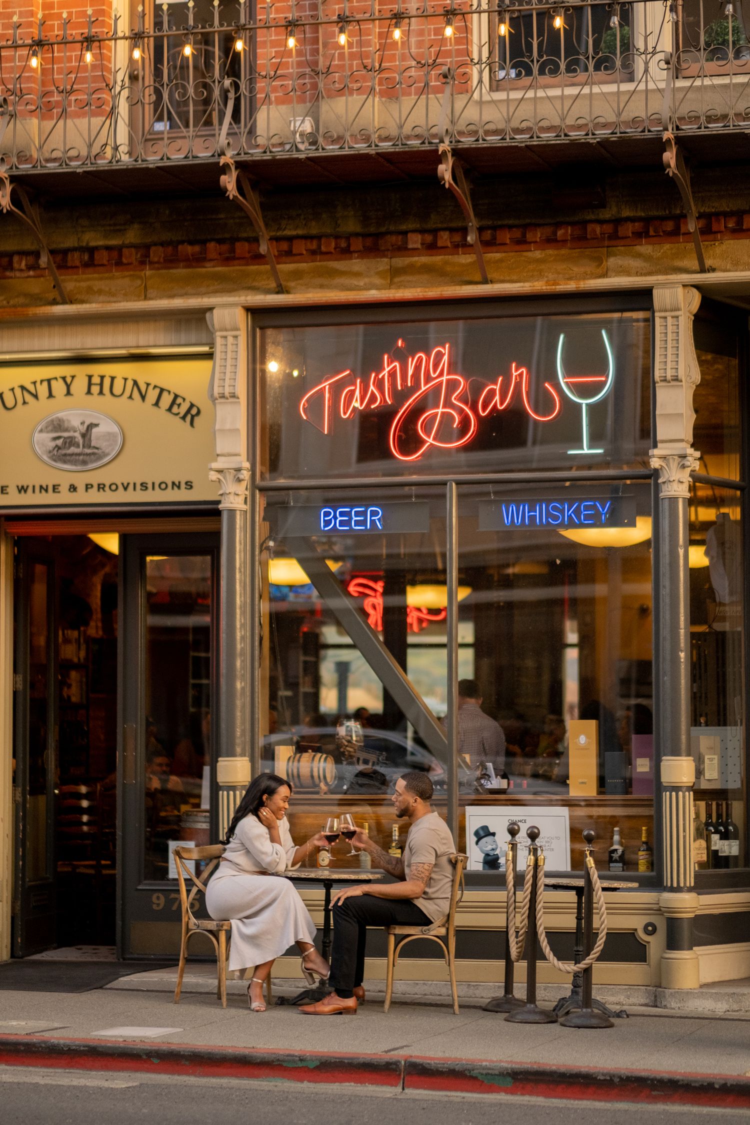An engaged couple enjoys drinks at an outdoor table in front of a glowing Tasting Bar neon sign.
