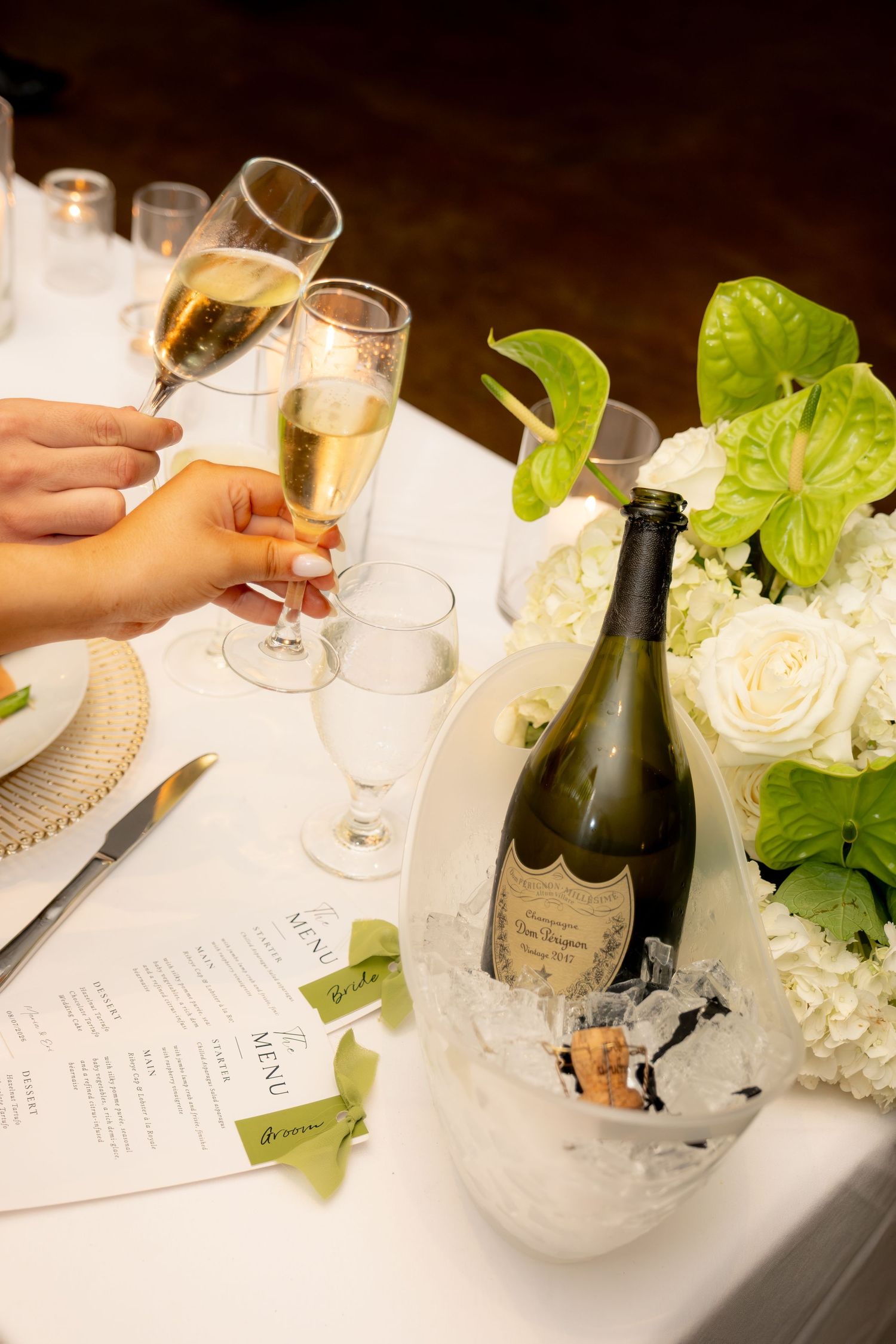 Hands toasting with champagne flutes beside Dom Perignon bottle and white floral centerpiece.