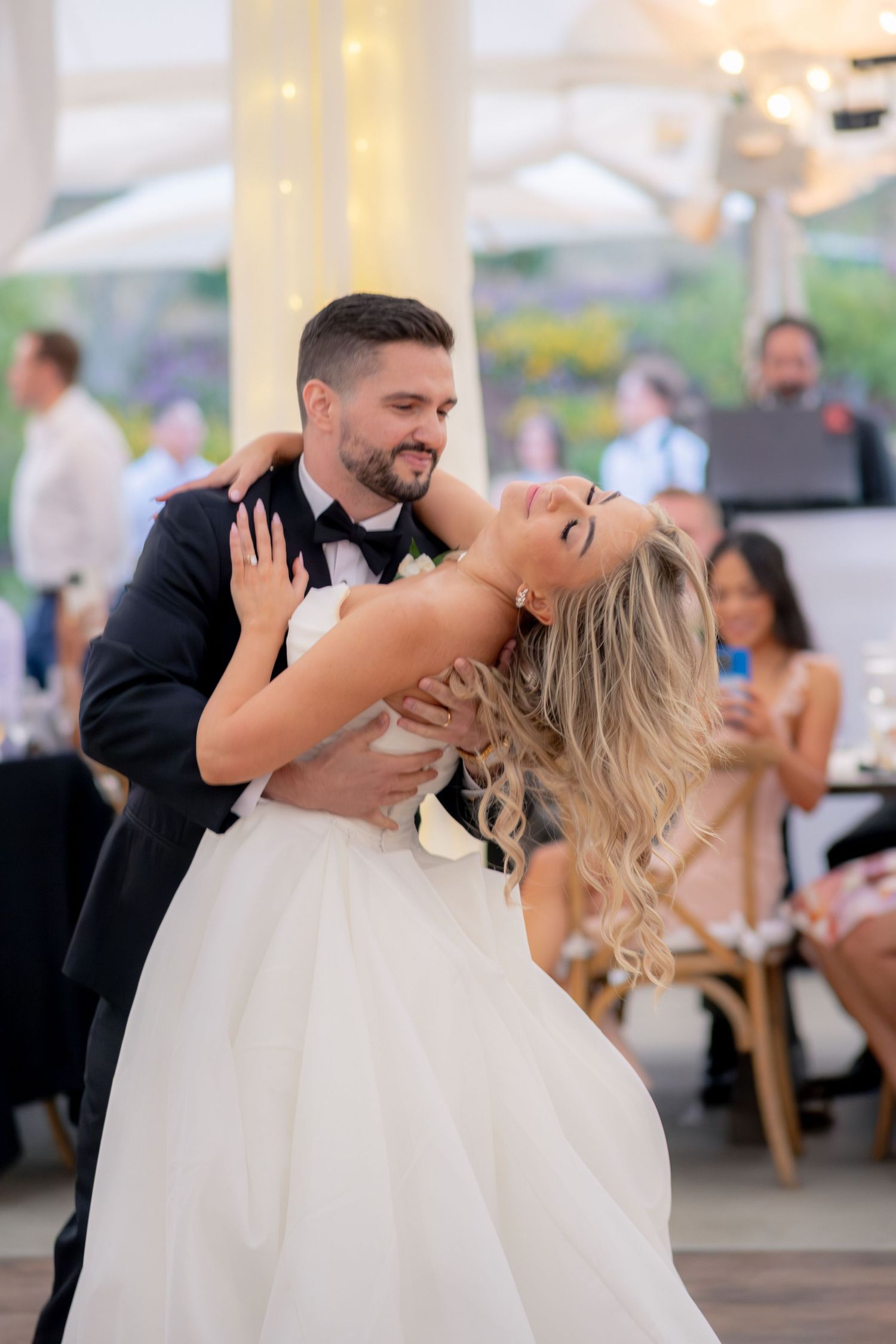 Groom dipping laughing bride during energetic first dance at outdoor wedding reception.