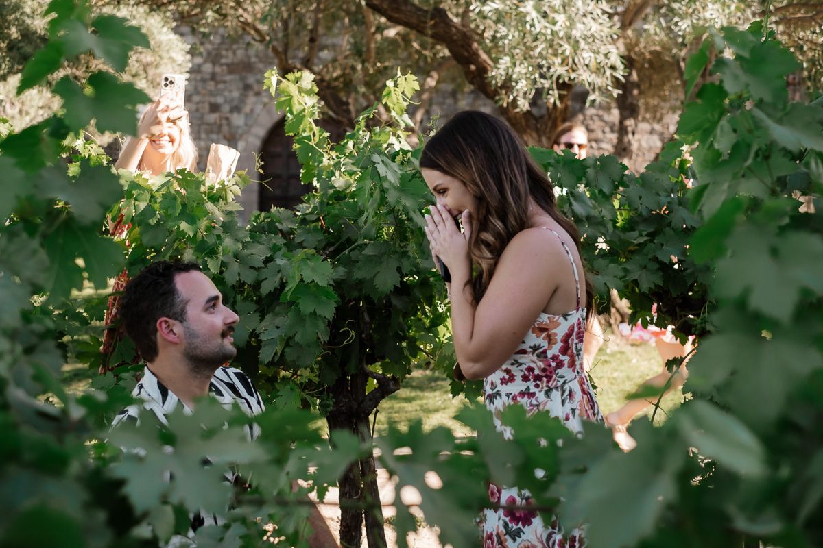 Man proposing on one knee to surprised woman covering her mouth in a lush vineyard setting.