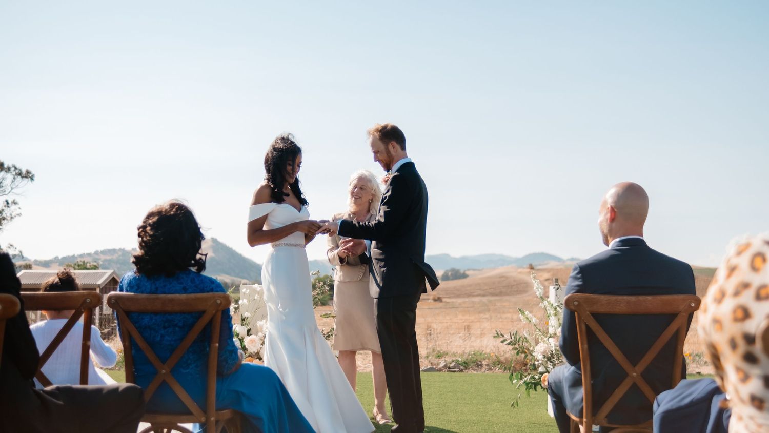 Couple holds hands during outdoor wedding ceremony with rolling hills backdrop.