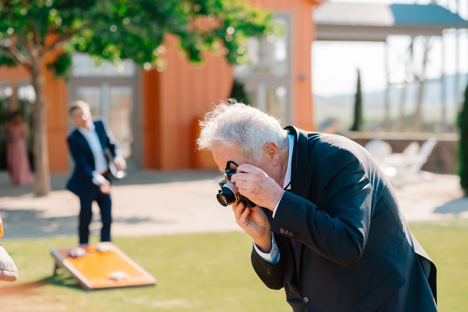 Elderly man photographs wedding guests playing cornhole at sunny outdoor reception venue.