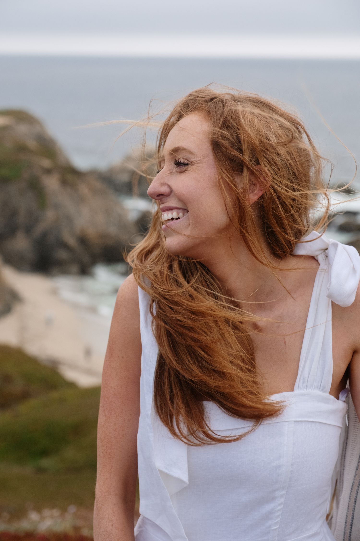 Happy young woman with long red hair laughing outdoors near rocky coastal cliffs with ocean in background.