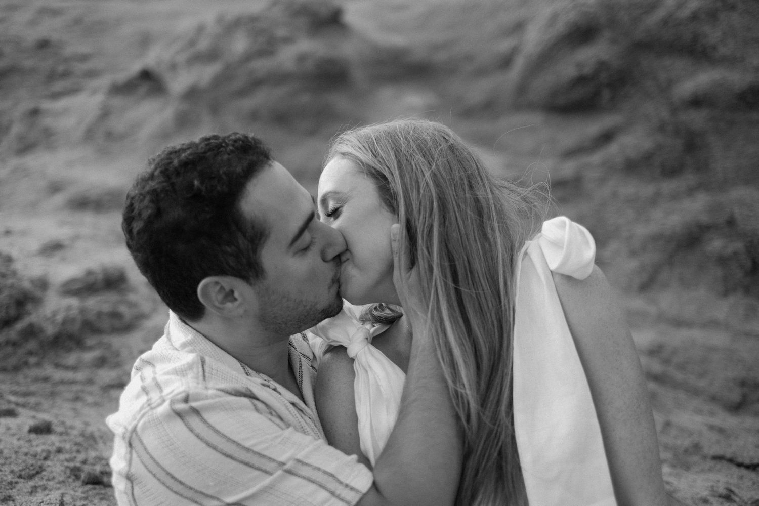 Black and white photo of couple sharing a passionate kiss on sandy beach.