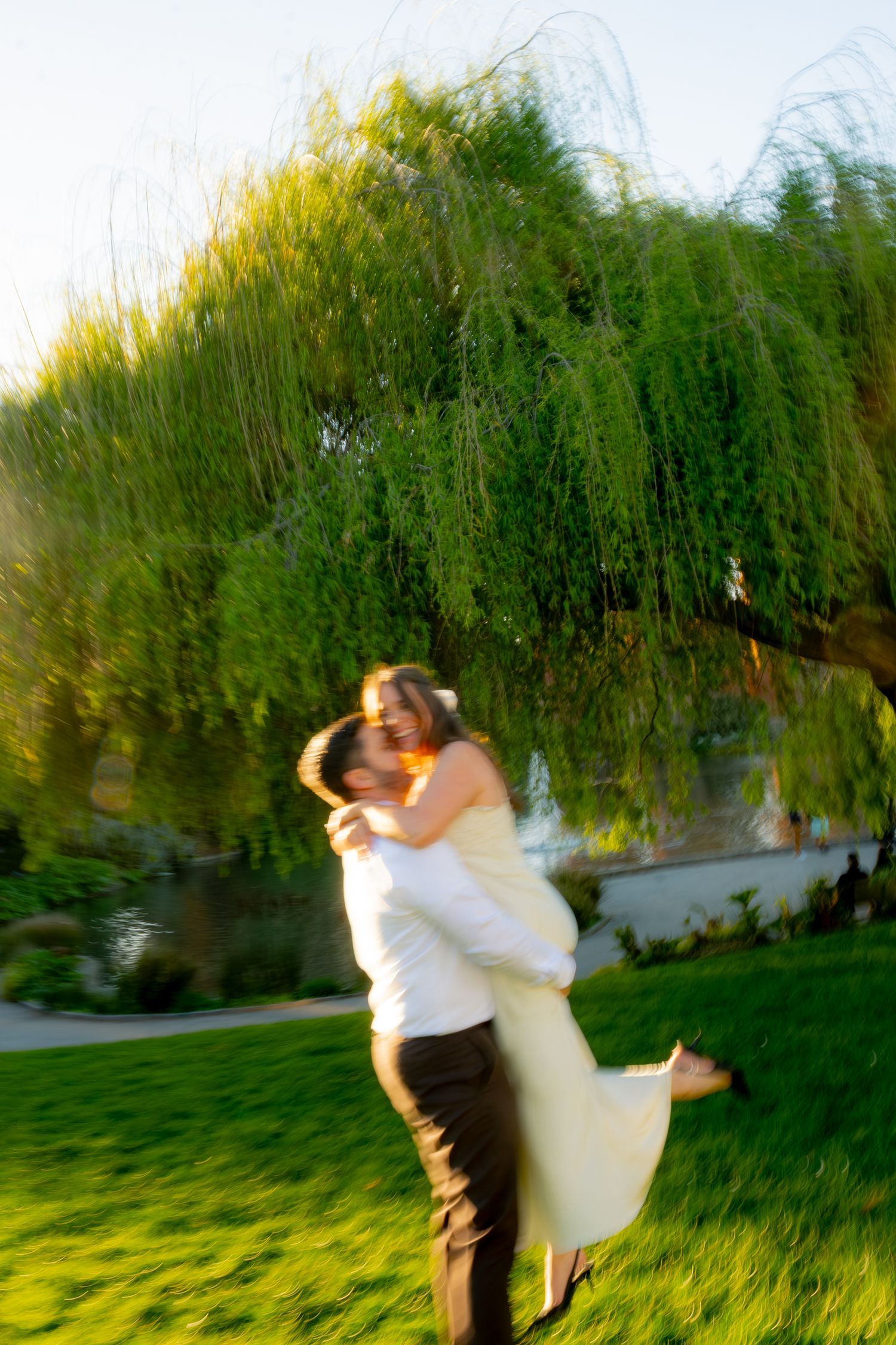 Groom lifting bride in white dress outdoors near weeping willow tree on green lawn during golden hour.