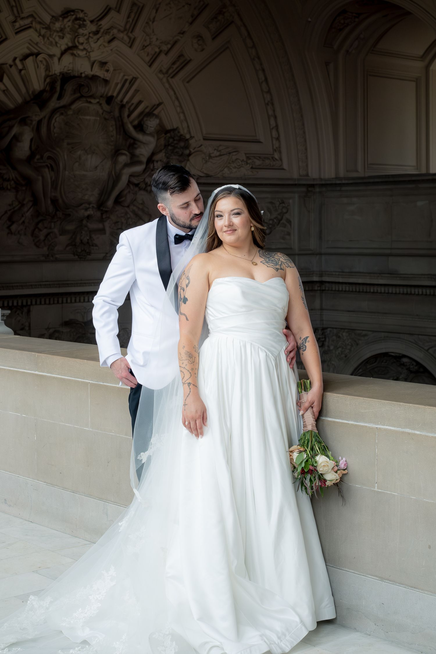 Wedding couple posing by carved stone wall, bride in strapless white gown holding bouquet, groom in white jacket.