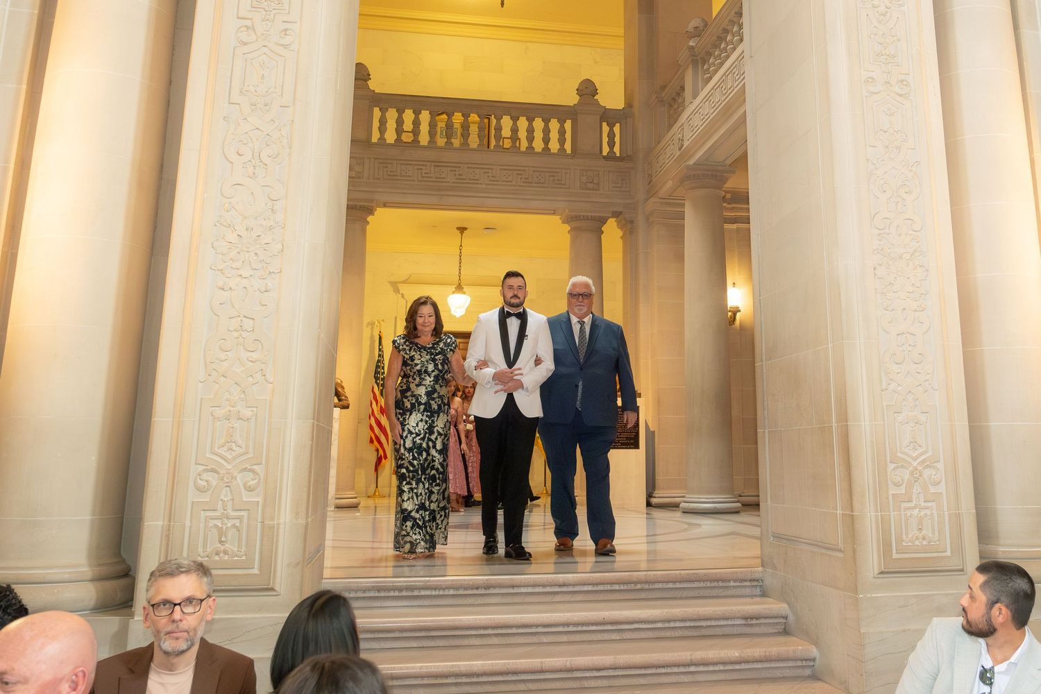 Three formally dressed individuals walk through an elegant columned hall with ornate architecture and marble stairs.
